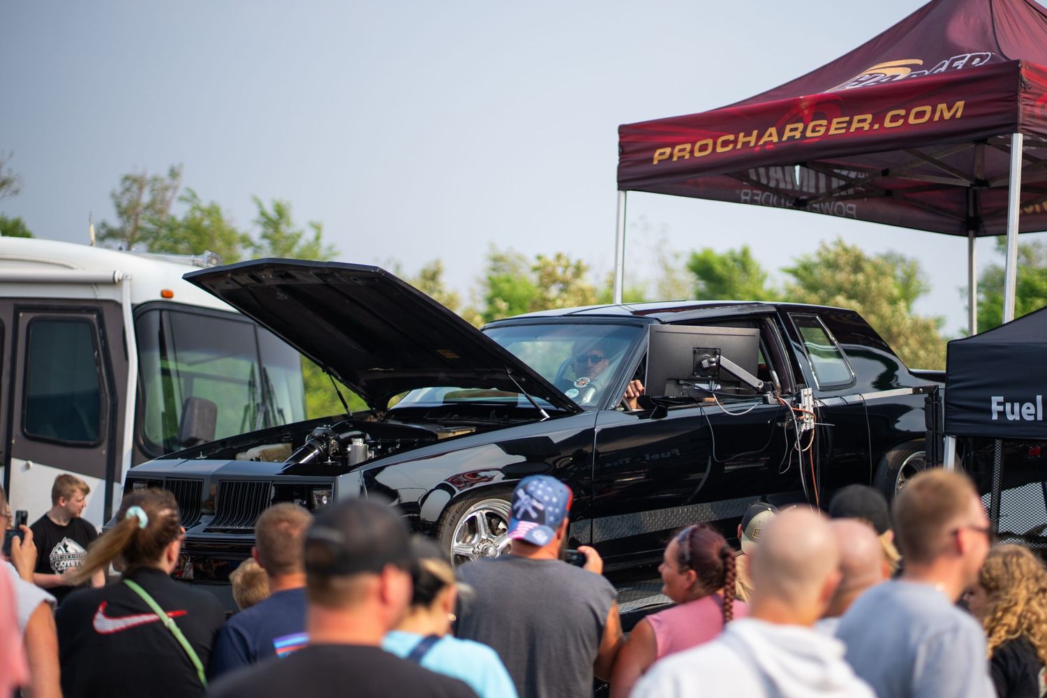 Crowd gathered at an outdoor automotive event with vehicles and tents visible.