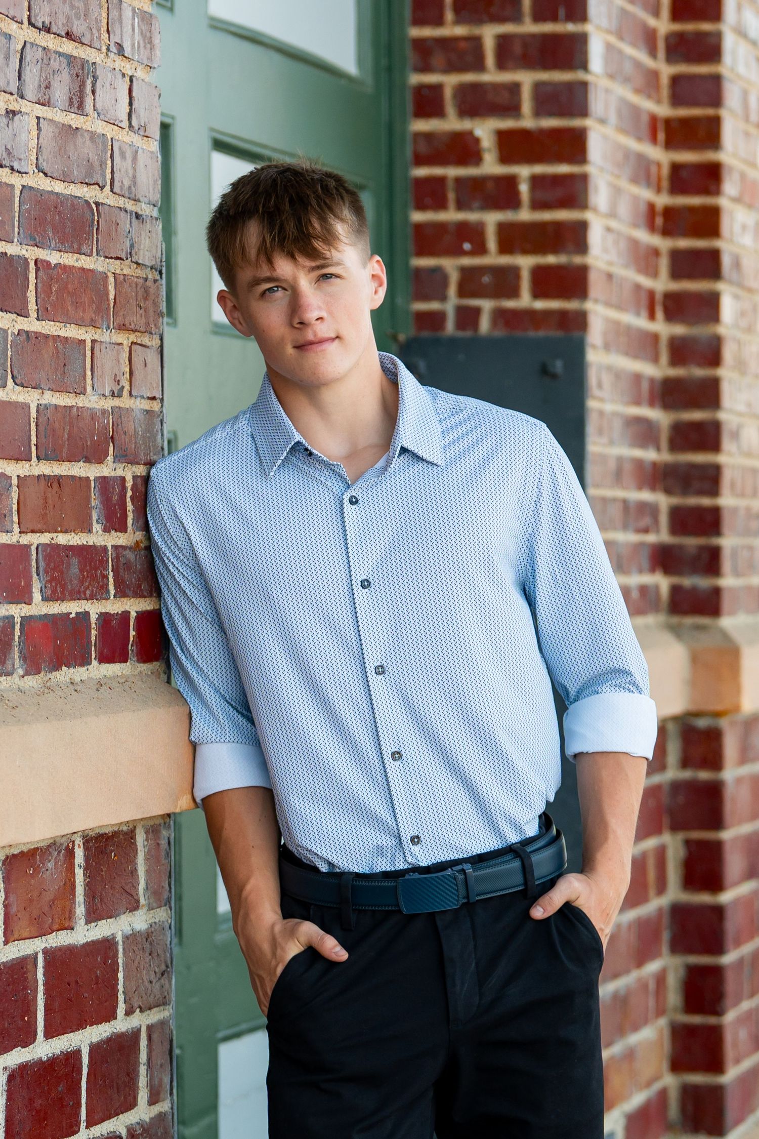 A relaxed pose against brick wall wearing business casual attire with hands in pockets.