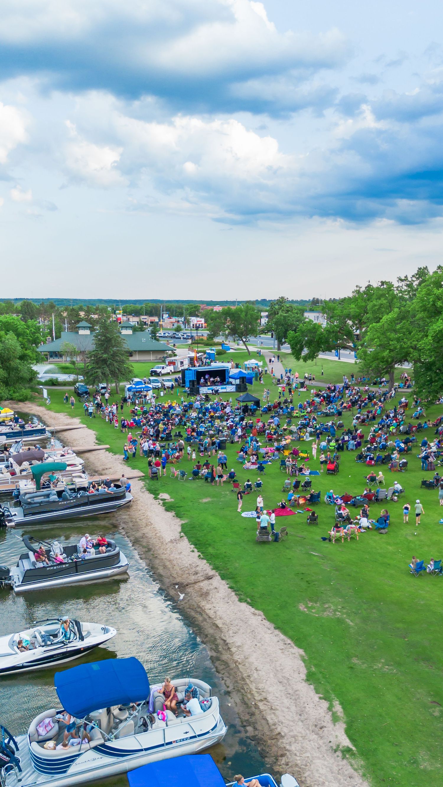 Aerial view of crowded lakeside event with boats and people on grass.