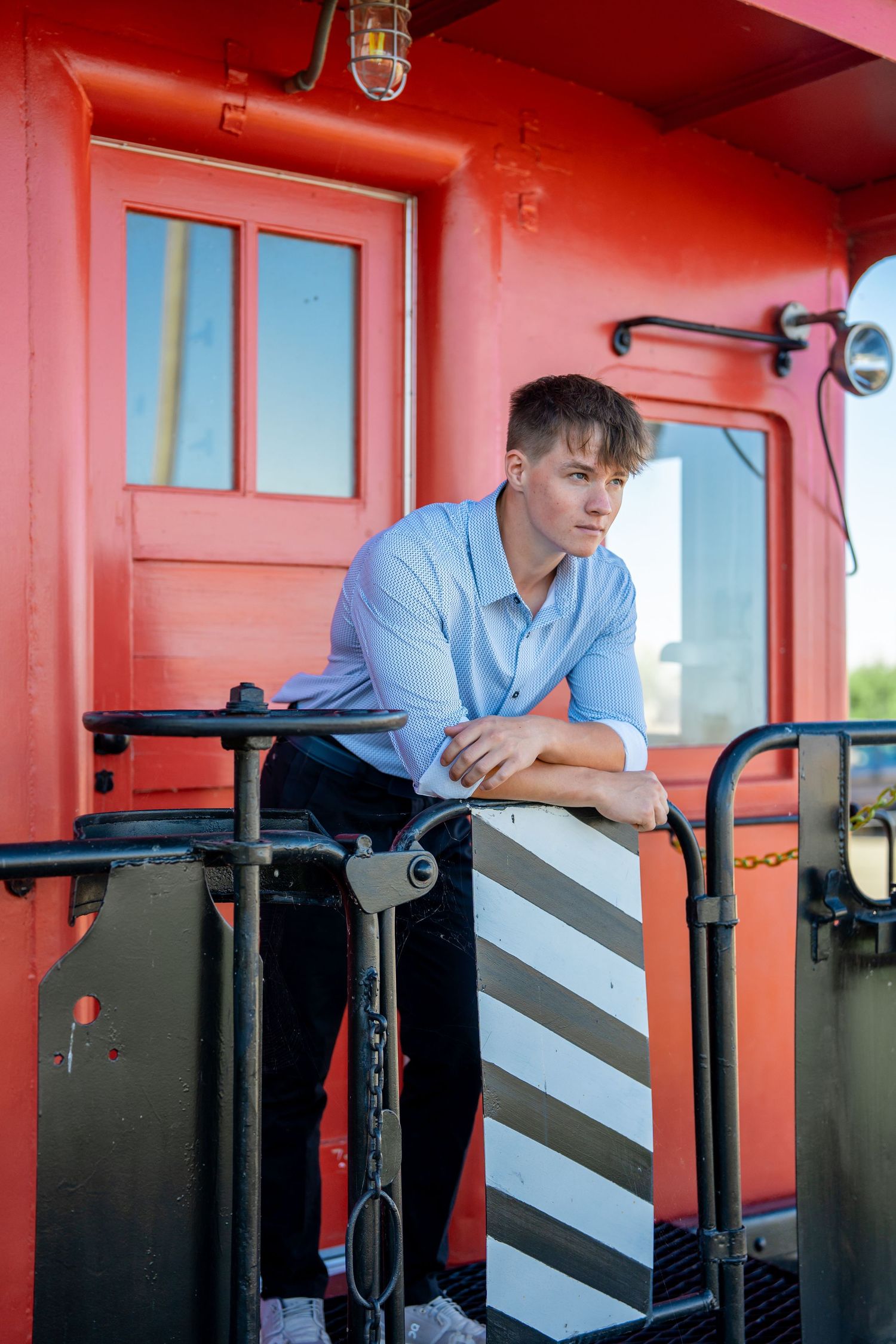 A person in light blue shirt leaning on railing of red painted train car exterior.