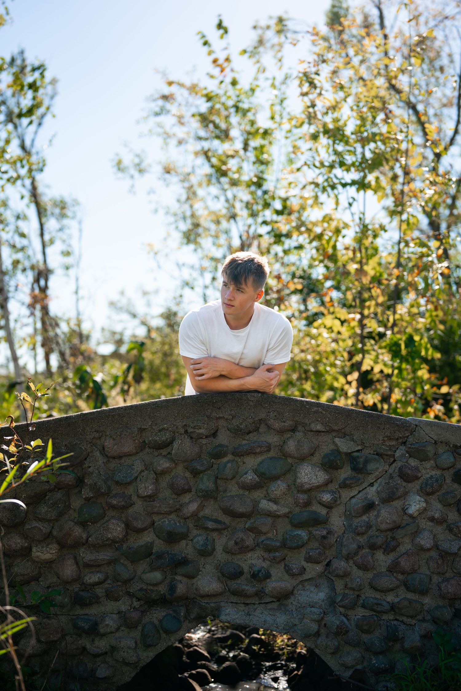 Person in white t-shirt leaning over a rustic stone wall surrounded by autumn foliage.