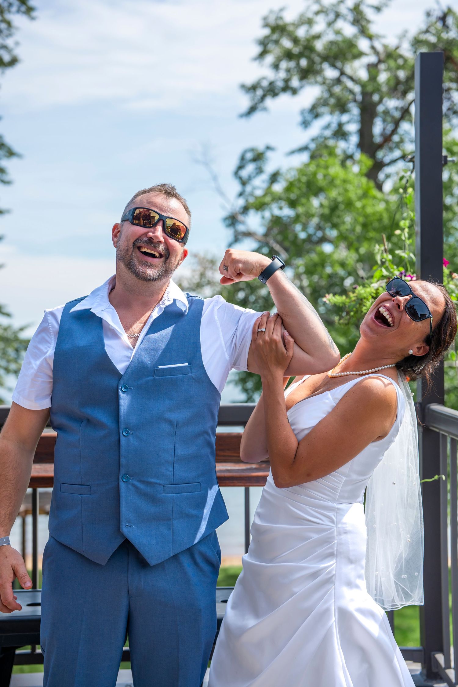 Newlyweds laugh and dance together on a sunny deck, with the groom in a blue vest and bride in white.