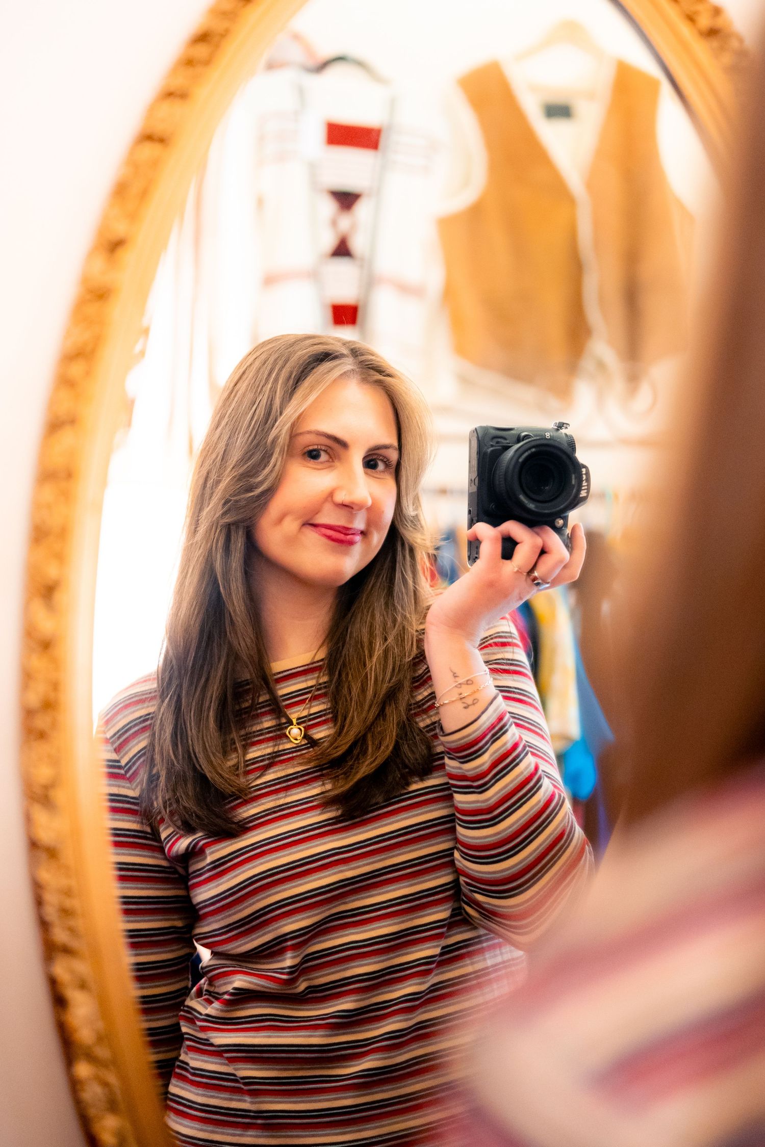 Woman in striped top taking mirror selfie with black camera in gold oval mirror, clothes visible in background.