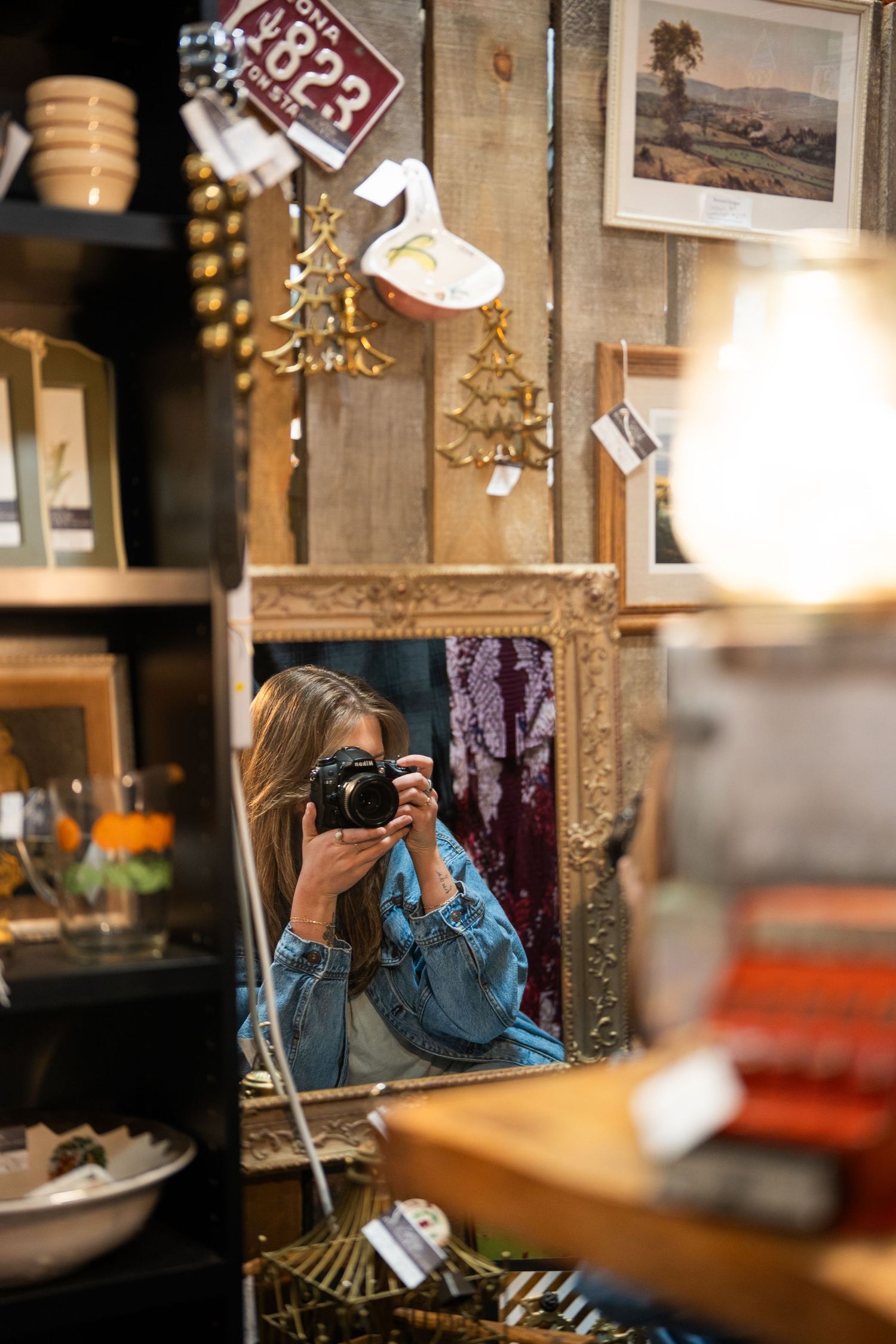 Woman taking mirror selfie with camera in eclectic antique shop with vintage frames and decorative items on shelves.