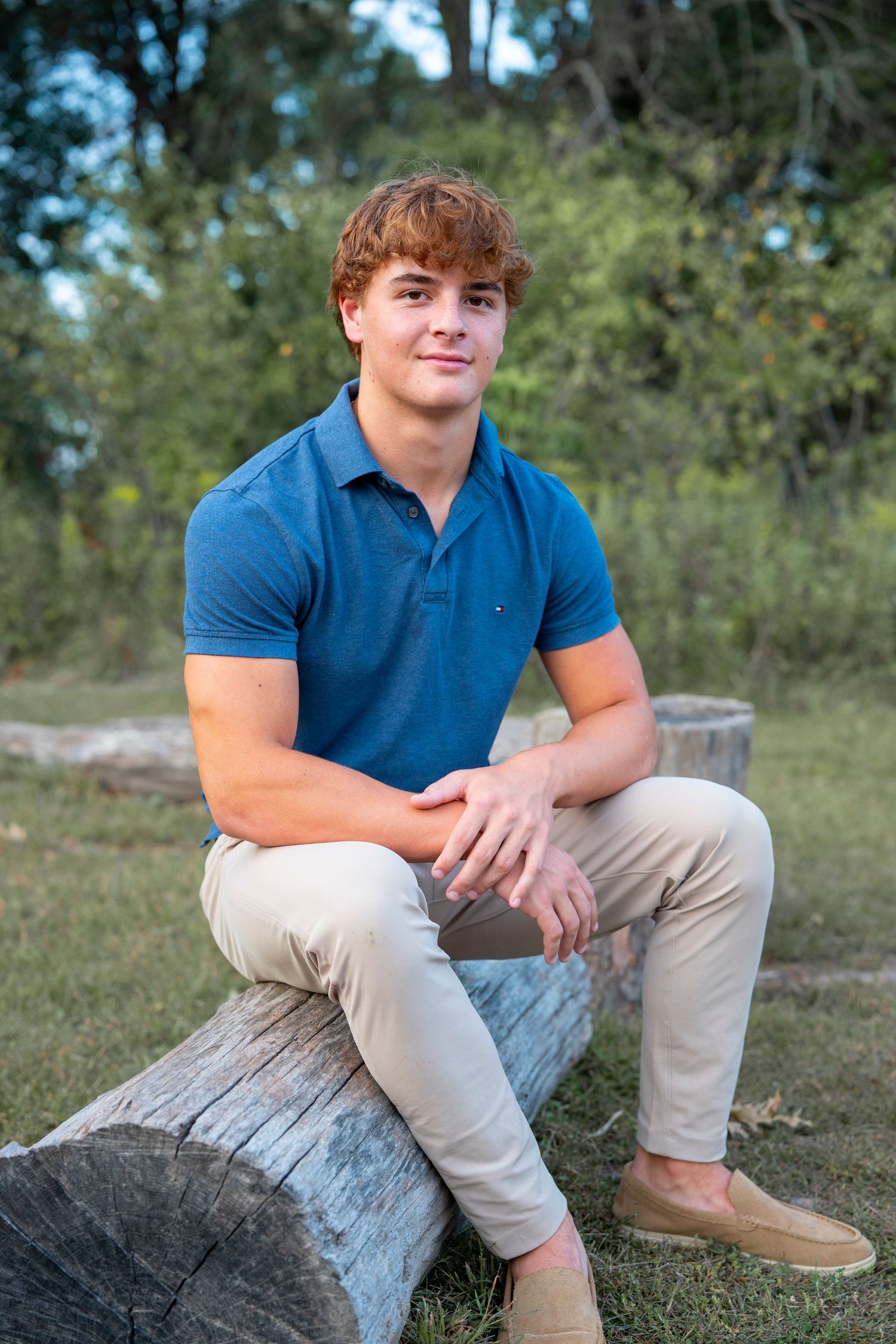 Candid outdoor senior photo showing someone seated on a wooden log in casual attire.
