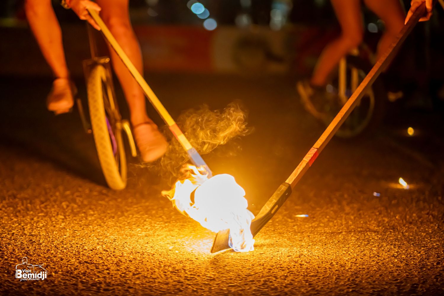 Flaming unicycle hockey stick illuminates dark pavement with bright orange glow during nighttime performance.