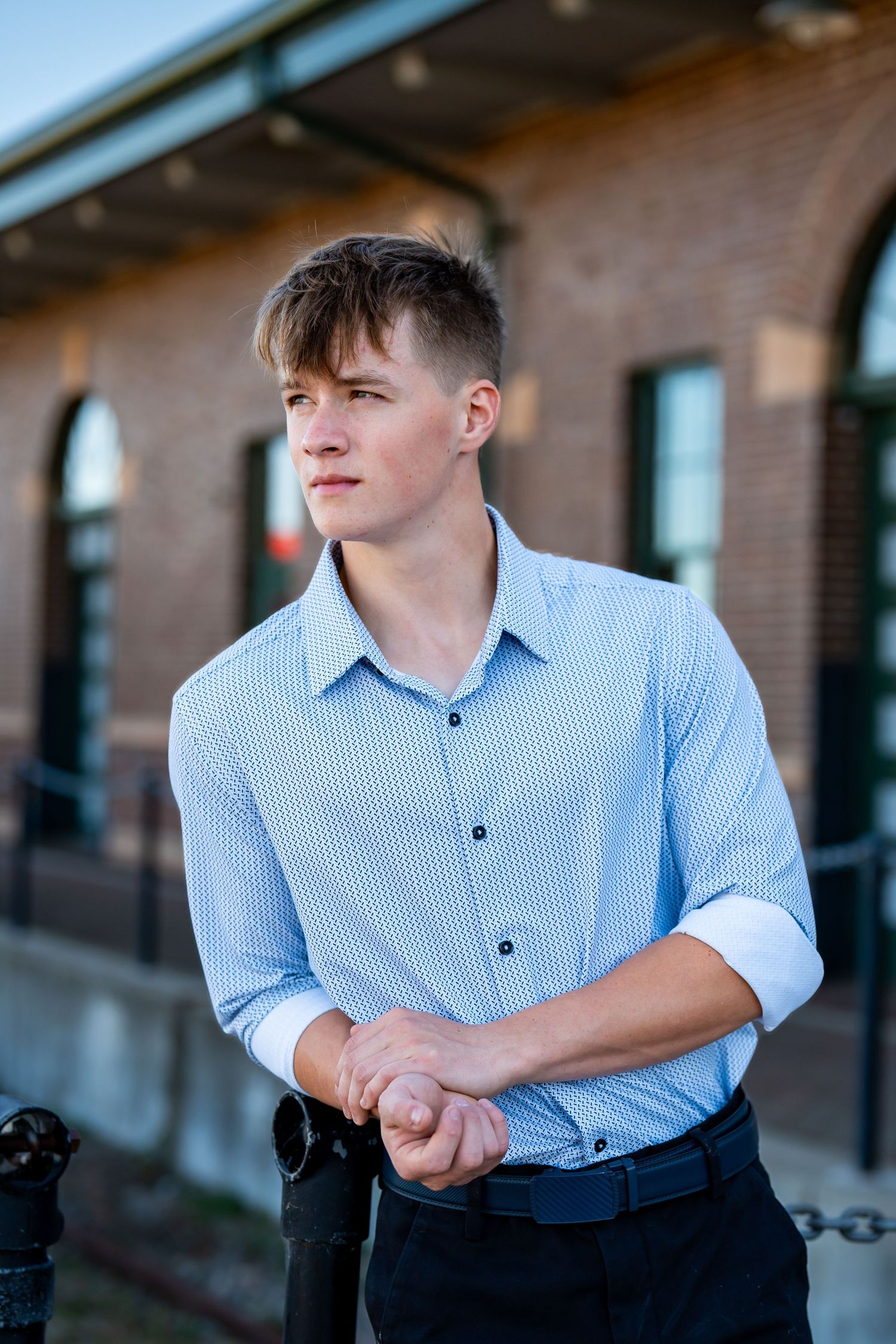 Person in light blue striped shirt and dark pants poses outdoors against brick building.