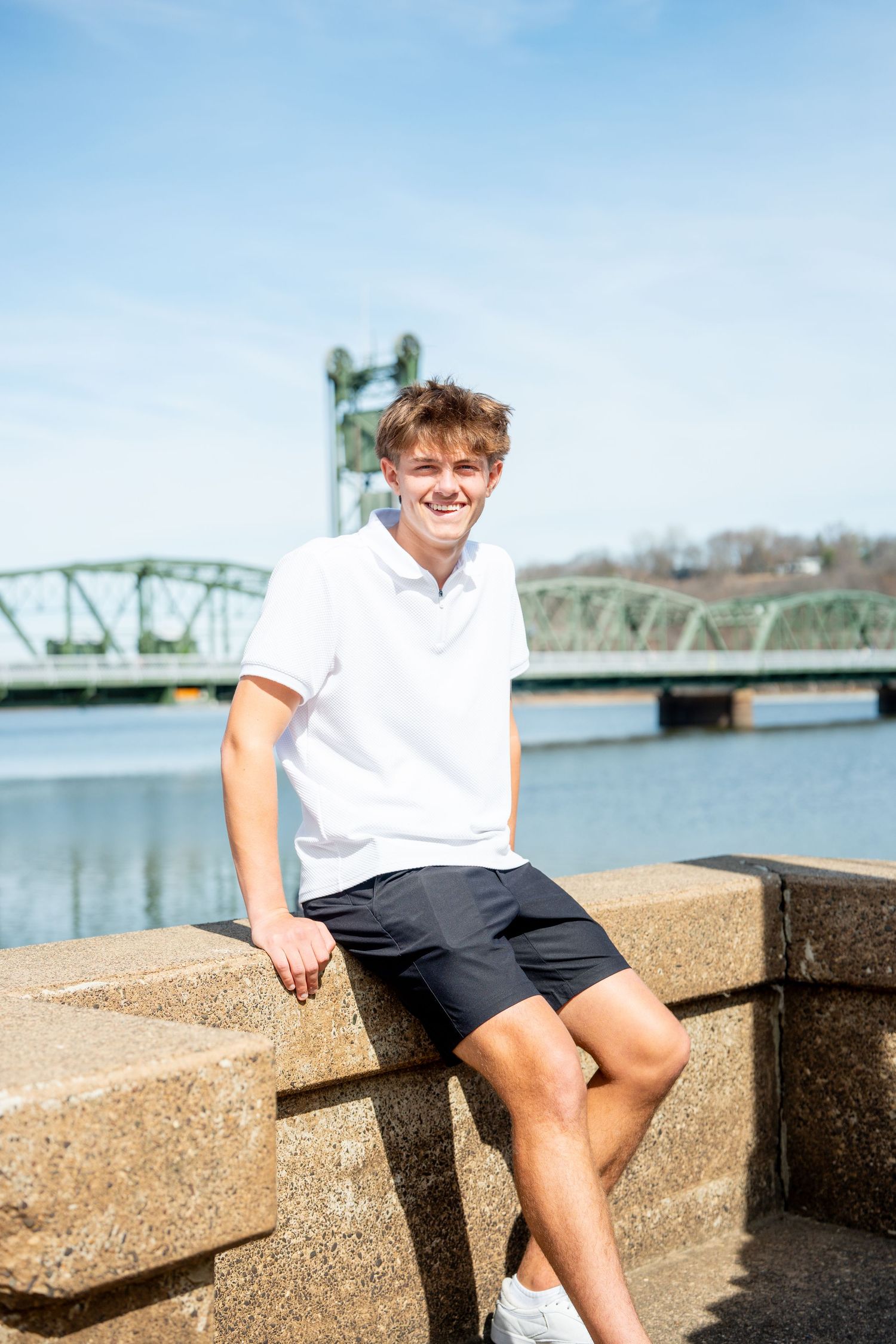 Person in white polo shirt and navy shorts sits on stone wall near green bridge over water on sunny day.