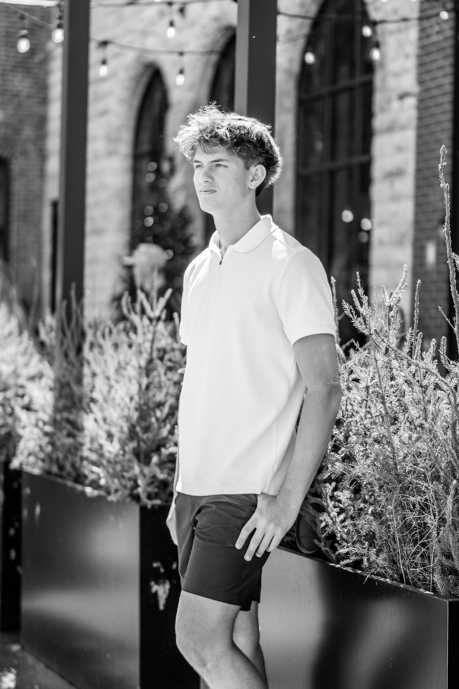 Black and white portrait of a young person in a white polo shirt and shorts standing beside flowering plants outdoors.