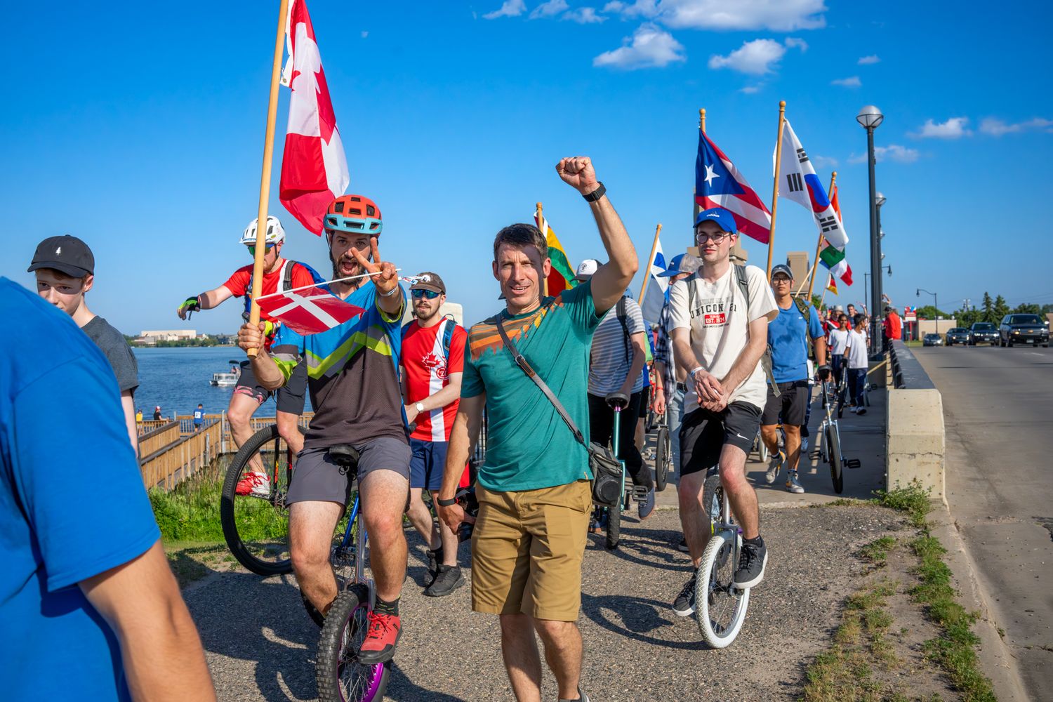 Group of unicyclists riding along waterfront path with flags waving in bright sunshine.