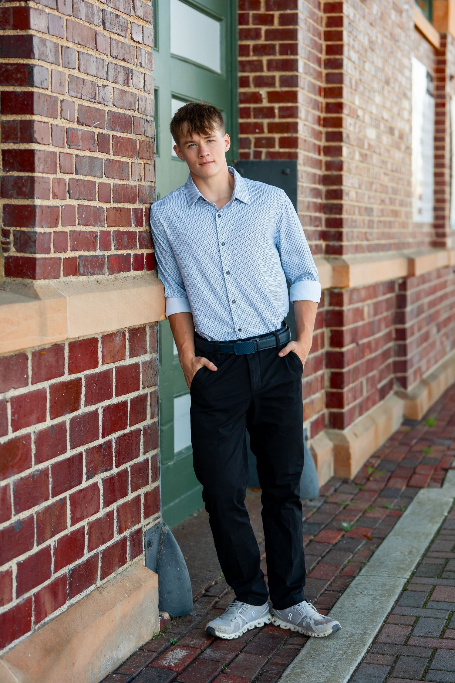 A casual portrait against brick wall wearing light blue shirt and dark pants with sneakers.