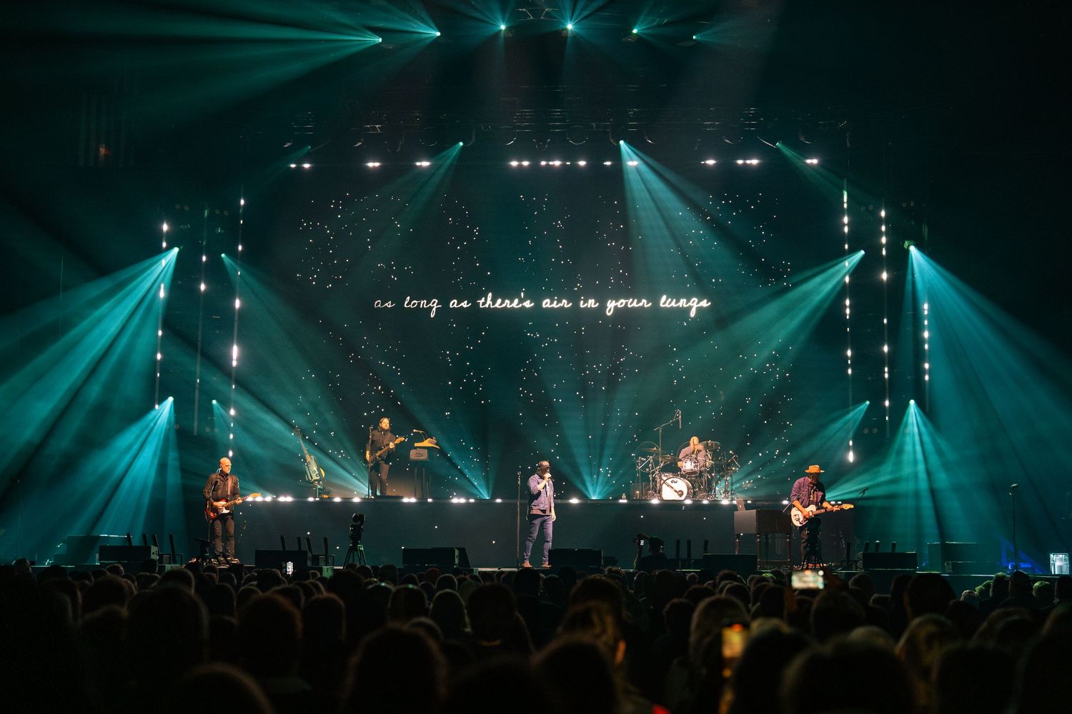 Wide concert venue with teal lighting and projection screens displaying worship lyrics.