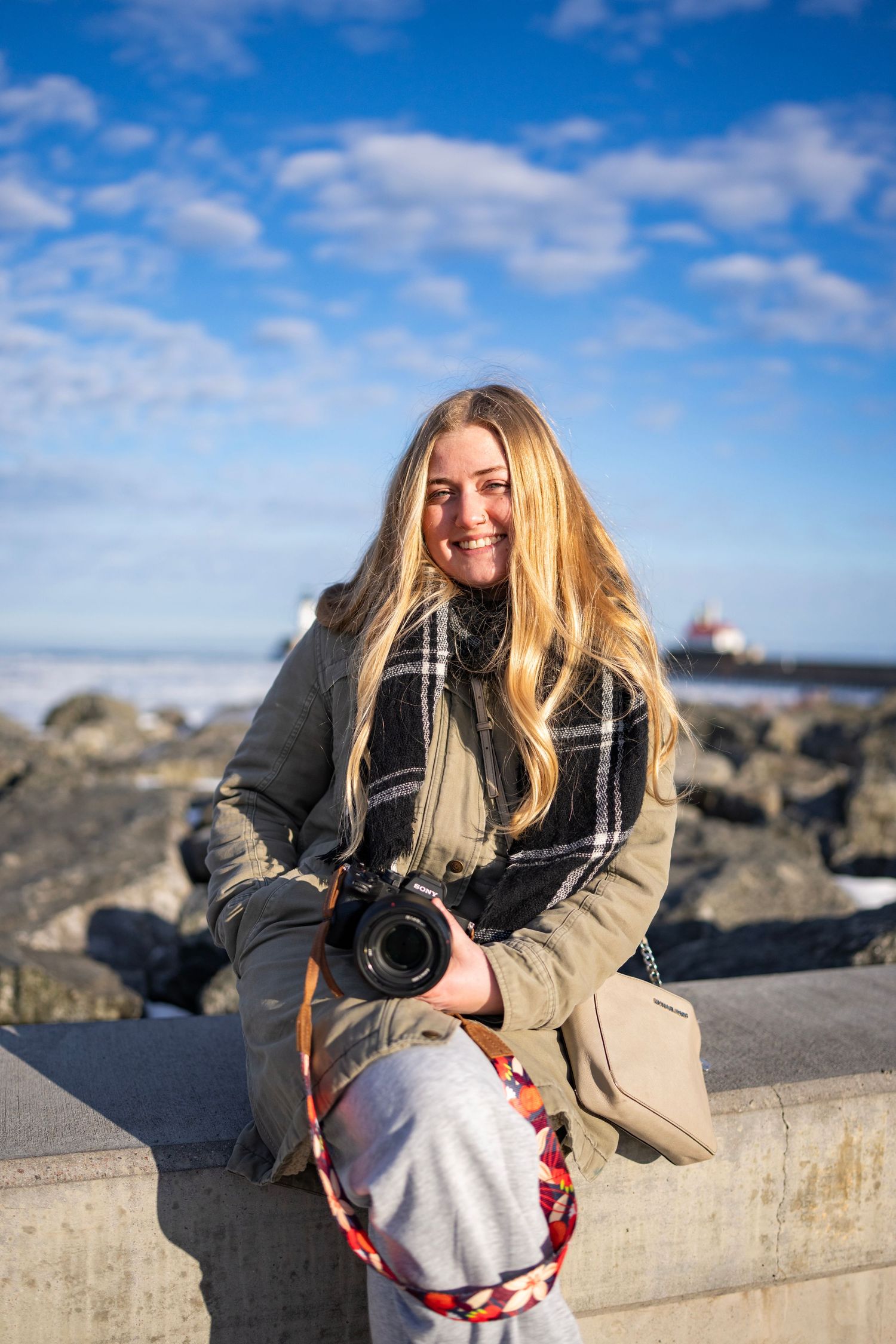 Smiling blonde woman holding a camera, wearing a plaid scarf and olive coat, sitting outdoors near rocky shoreline.
