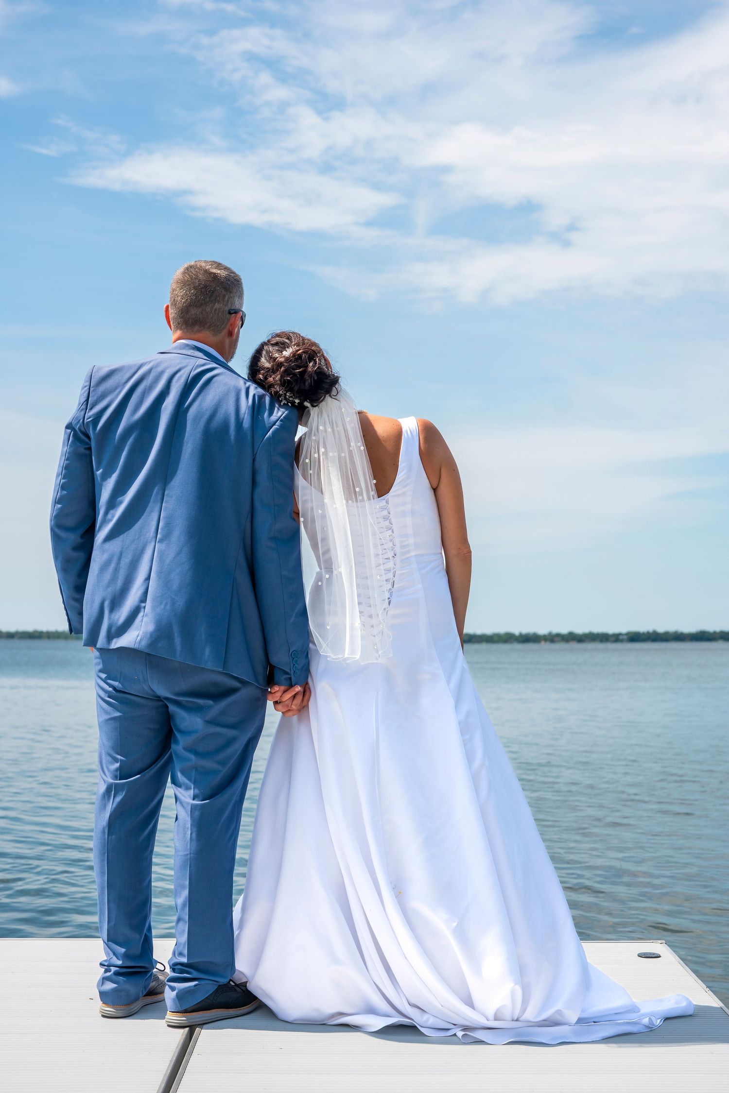 Bride in white dress and groom in blue suit sit together on dock edge with scenic lake view.