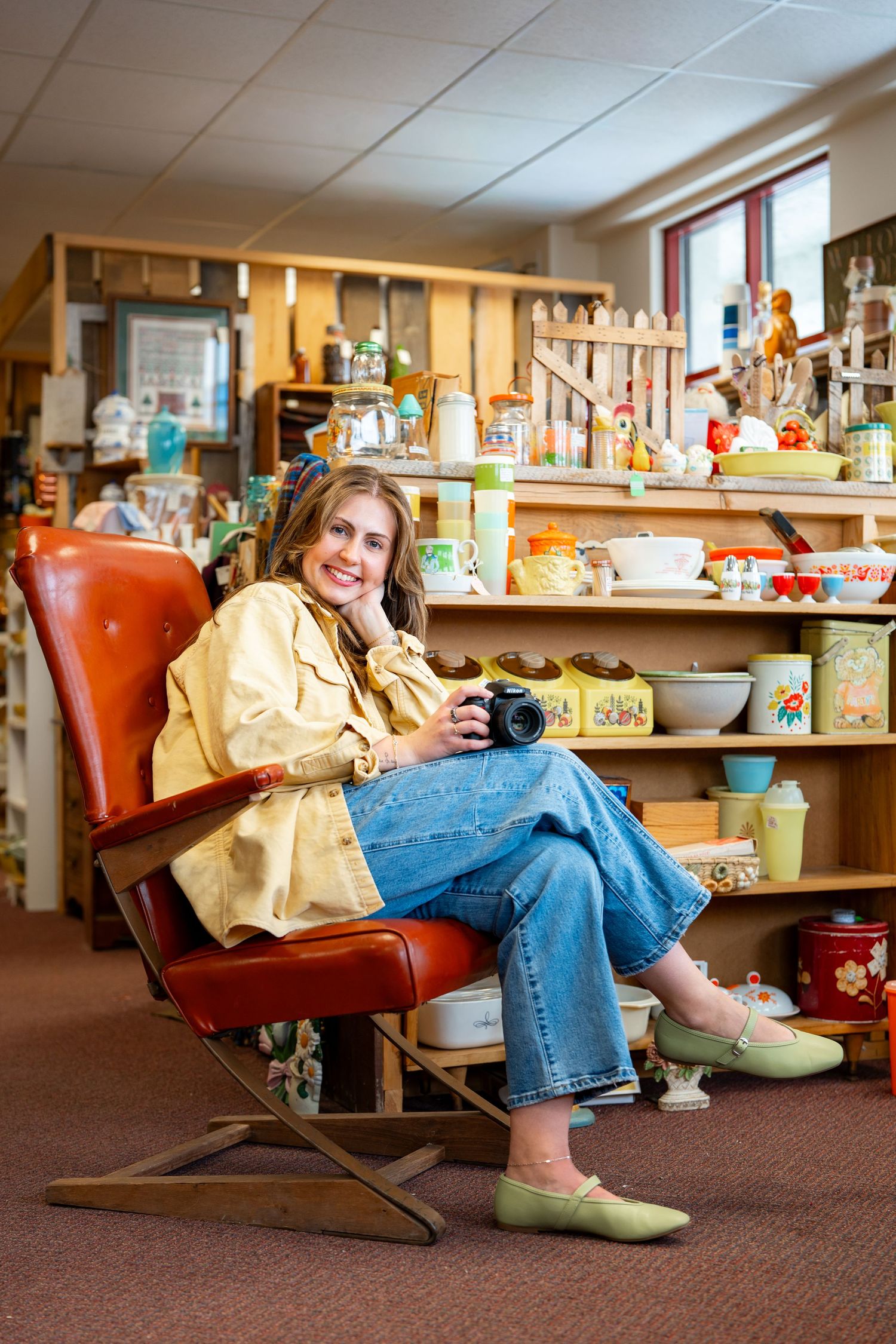 Woman sitting in a vintage red chair holding a camera, surrounded by colorful retro collectibles in an antique shop.