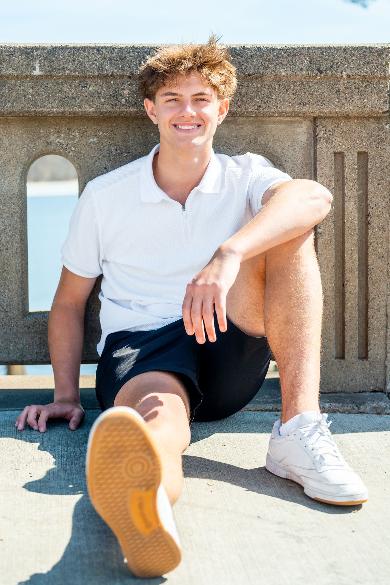 Person in white polo shirt and black pants sits casually on concrete ledge wearing white sneakers.