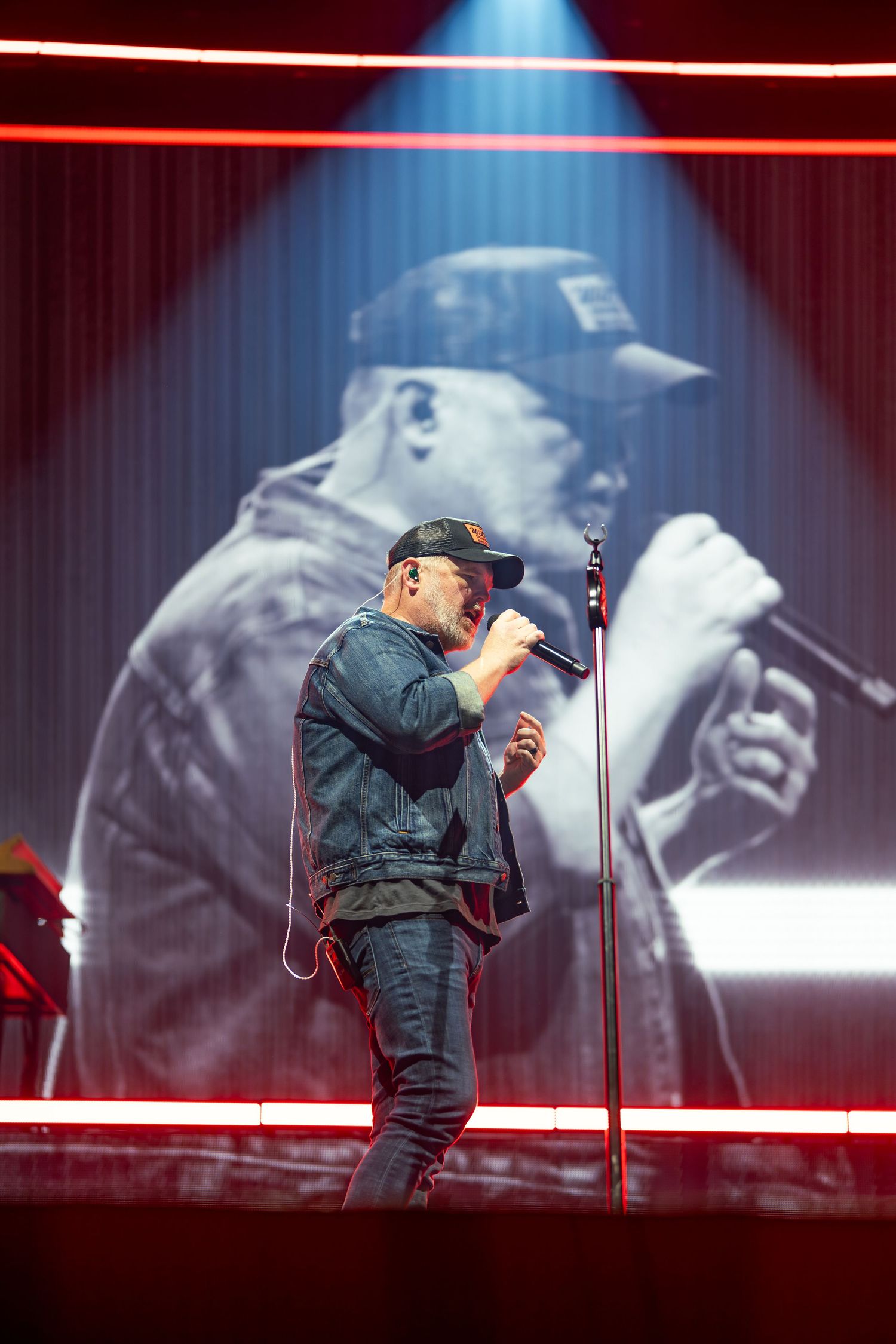 A performer in dark clothing stands on stage with dramatic backlighting and large video screens.
