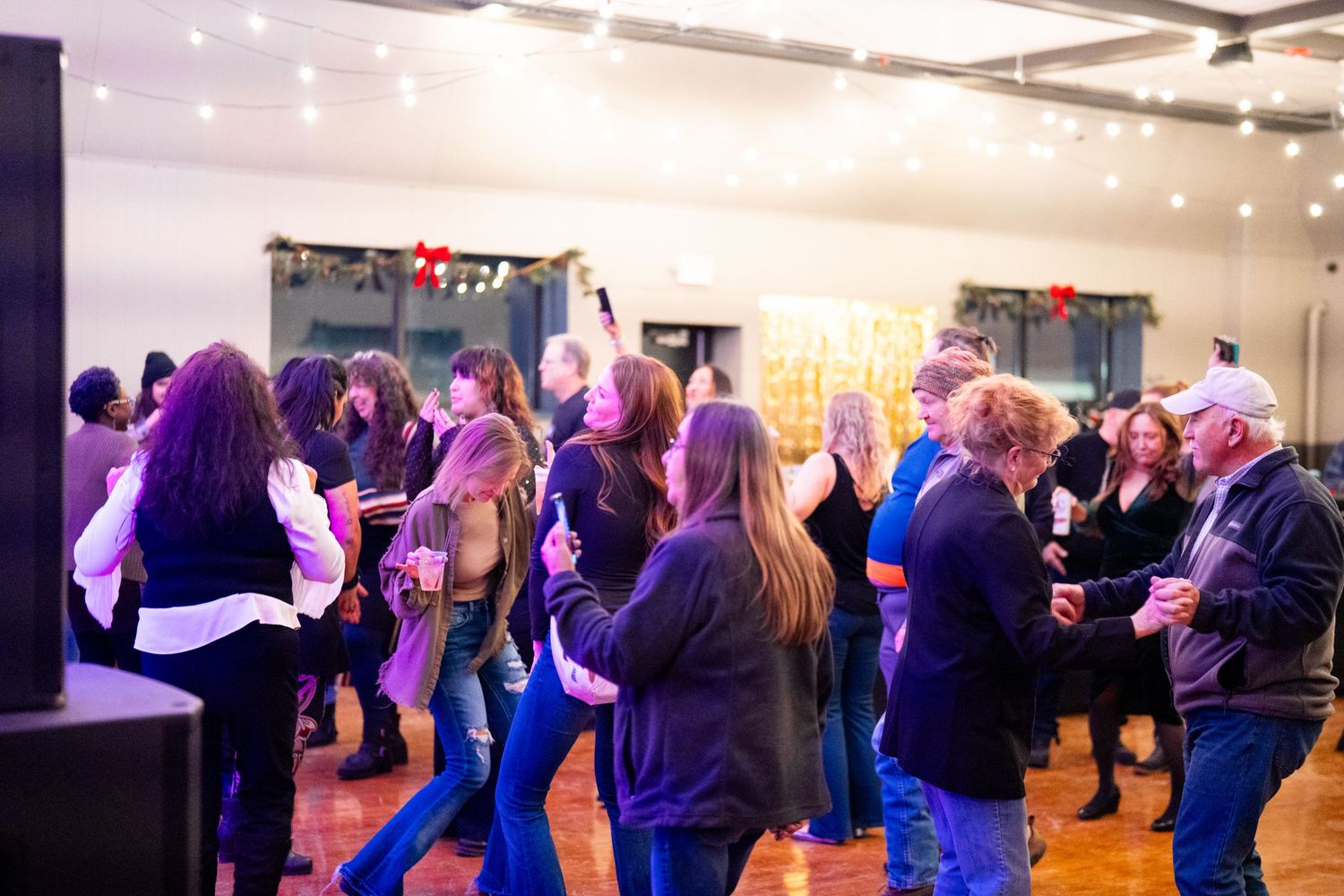 Crowd of people mingling and dancing at an indoor event with purple lighting and string lights.