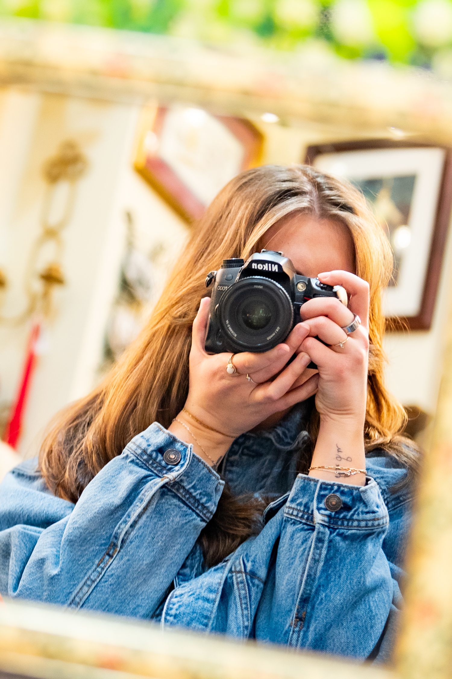 Young woman in denim jacket taking a mirror selfie with a Nikon DSLR camera in a vintage shop.