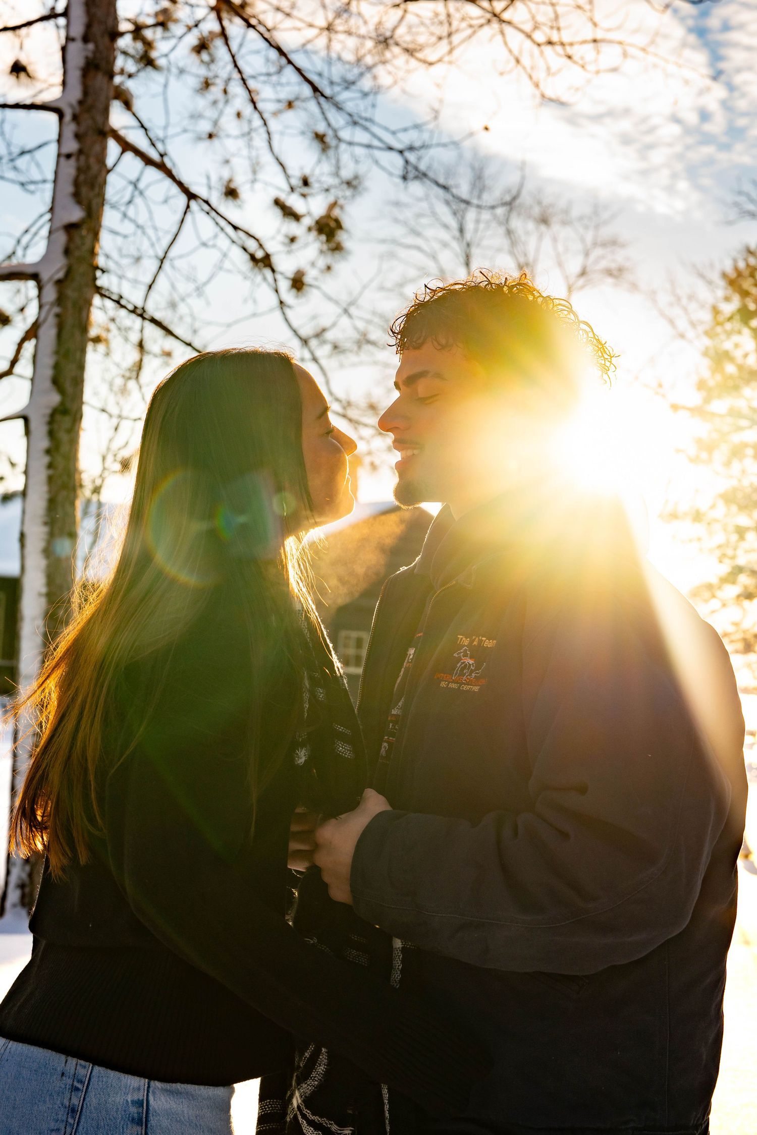 A silhouetted couple shares an intimate moment at sunset with sunlight streaming through bare winter trees.