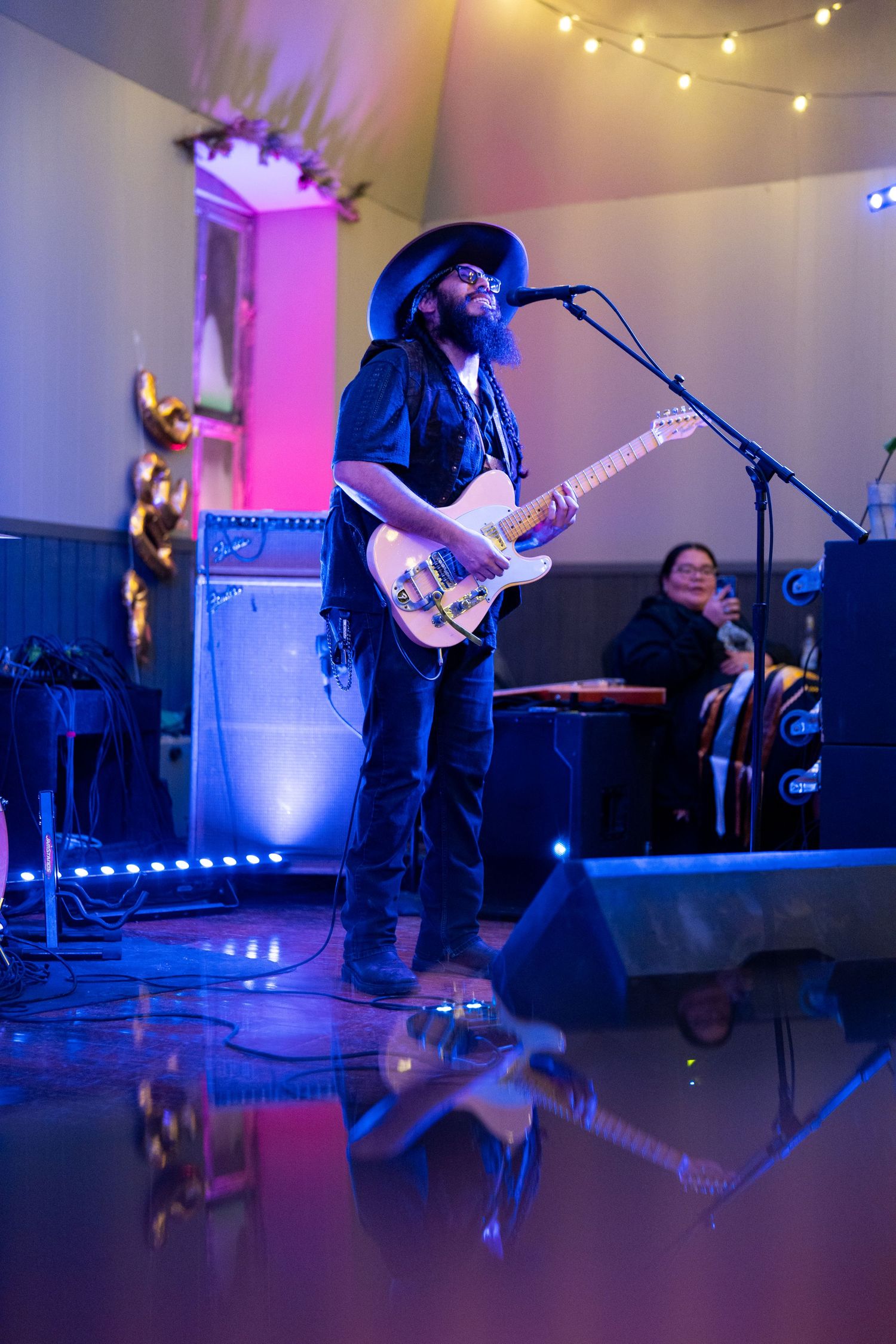 Musician playing guitar on stage with dramatic purple lighting and string light decorations.
