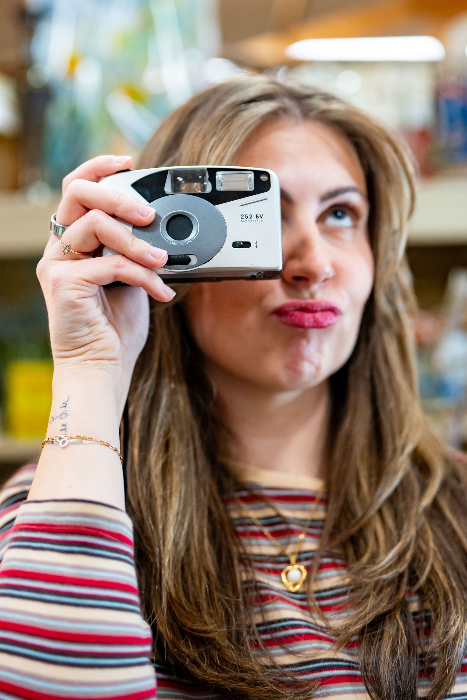 Young woman with long brown hair holding a vintage point-and-shoot film camera up near her face, smiling playfully.