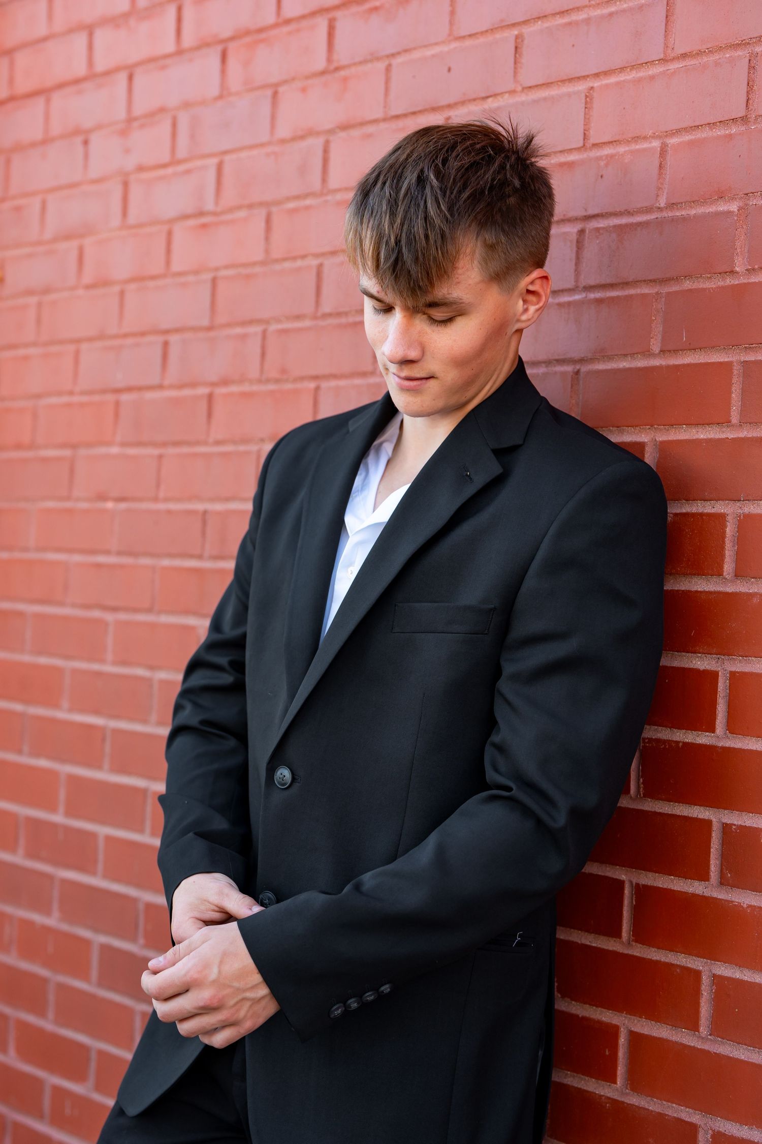 Person in black suit jacket leaning against red brick wall looking down.