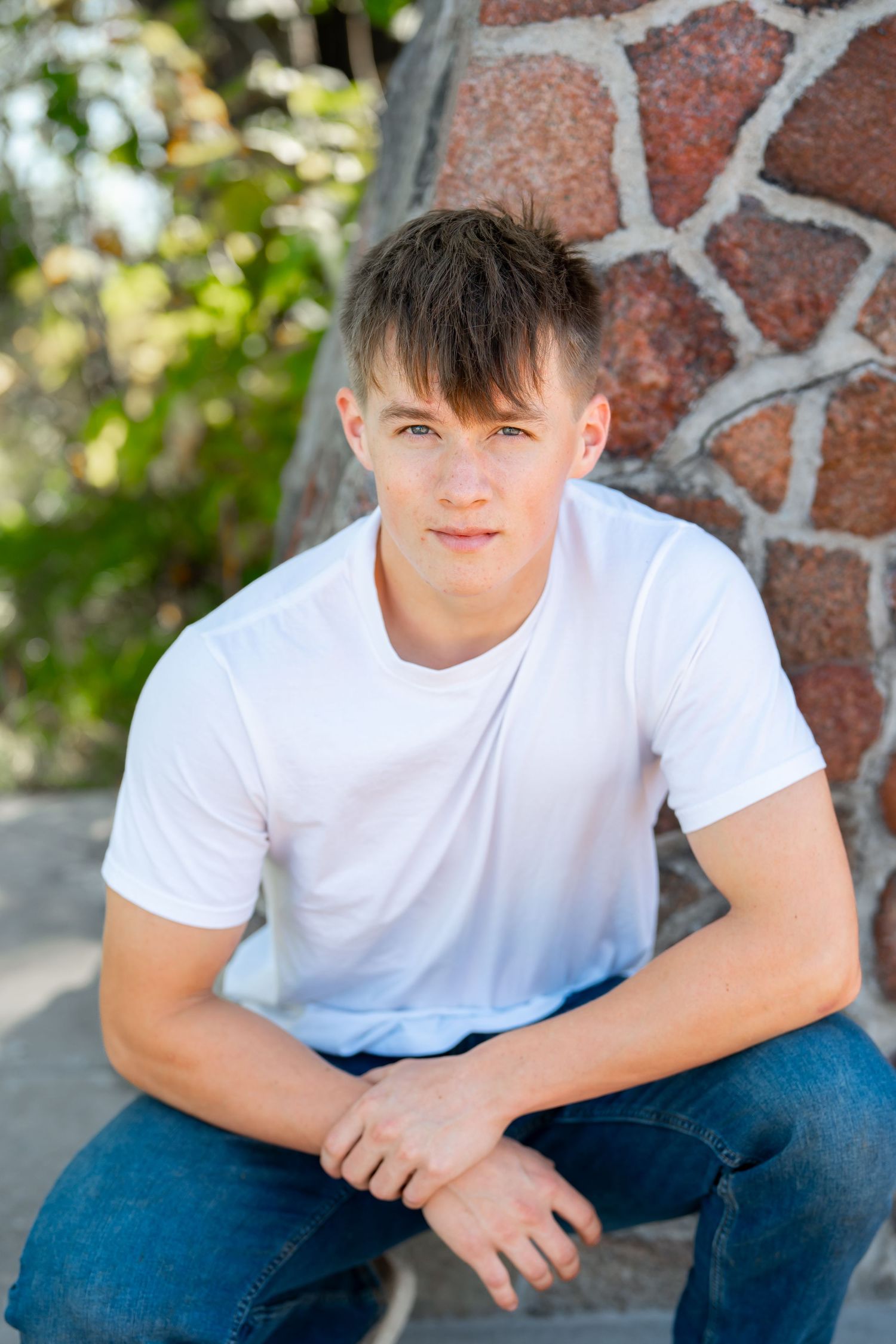 Casual portrait of young man in white t-shirt sitting by stone wall outdoors.