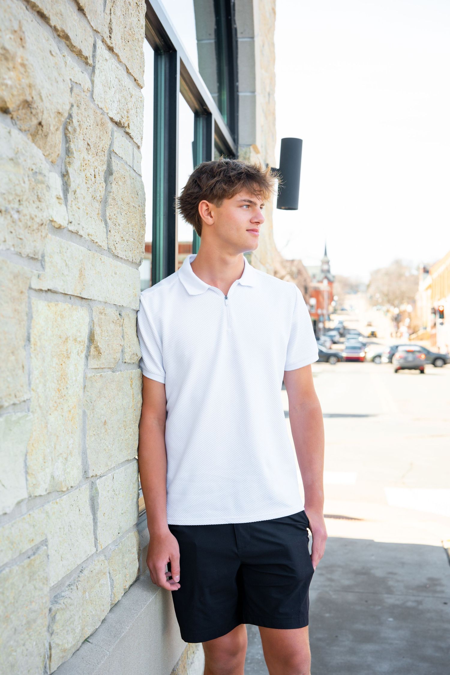 Person in white t-shirt and black shorts stands against stone wall in outdoor setting with cars visible in background.