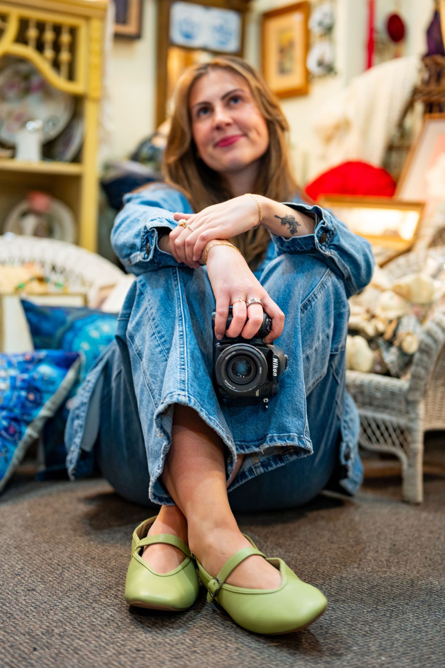 Woman in denim outfit sits on floor holding a camera, wearing green Mary Jane flats, surrounded by vintage decor.