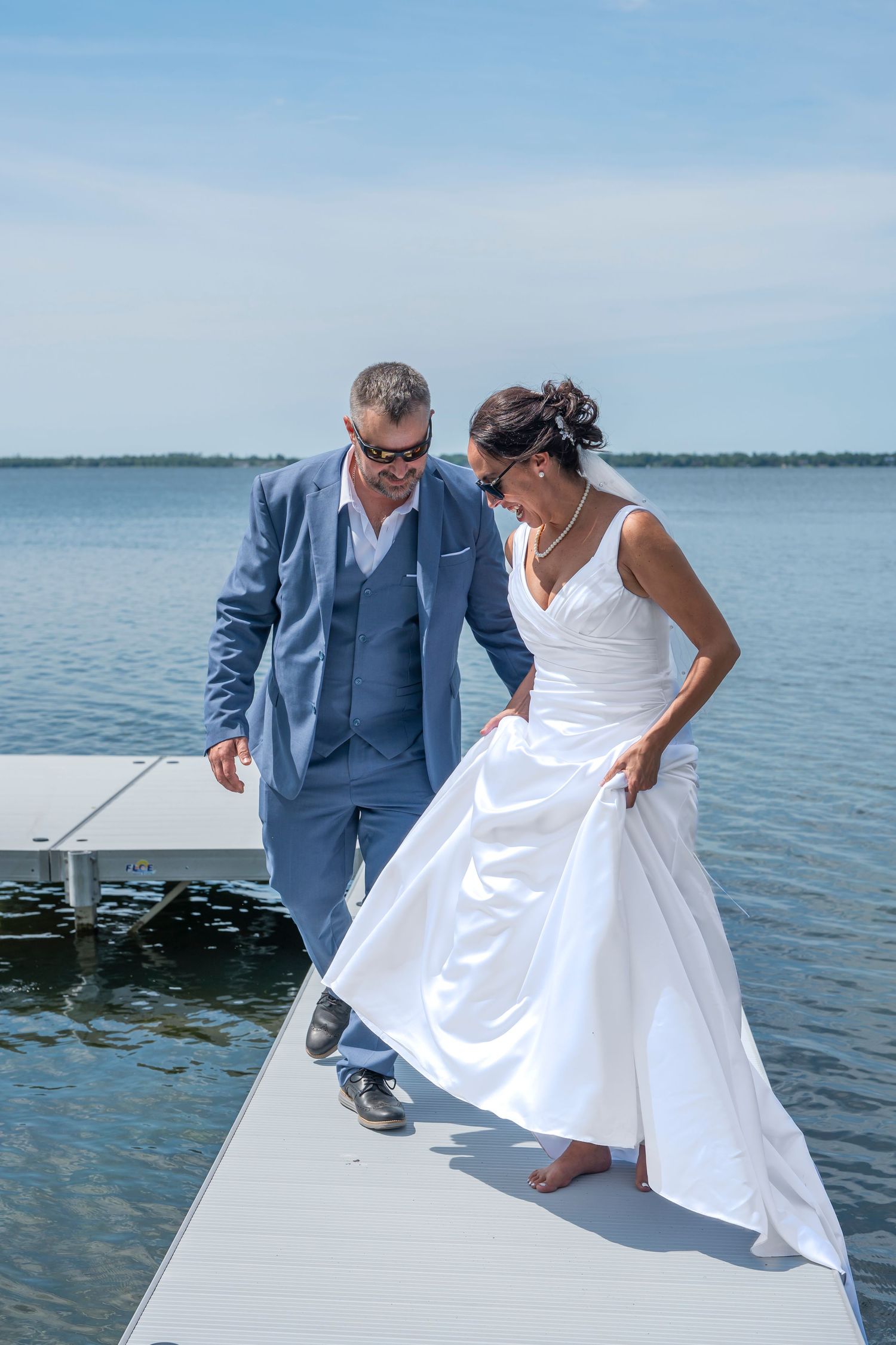 Couple in wedding attire playfully interacts on white dock platform over scenic waterfront.