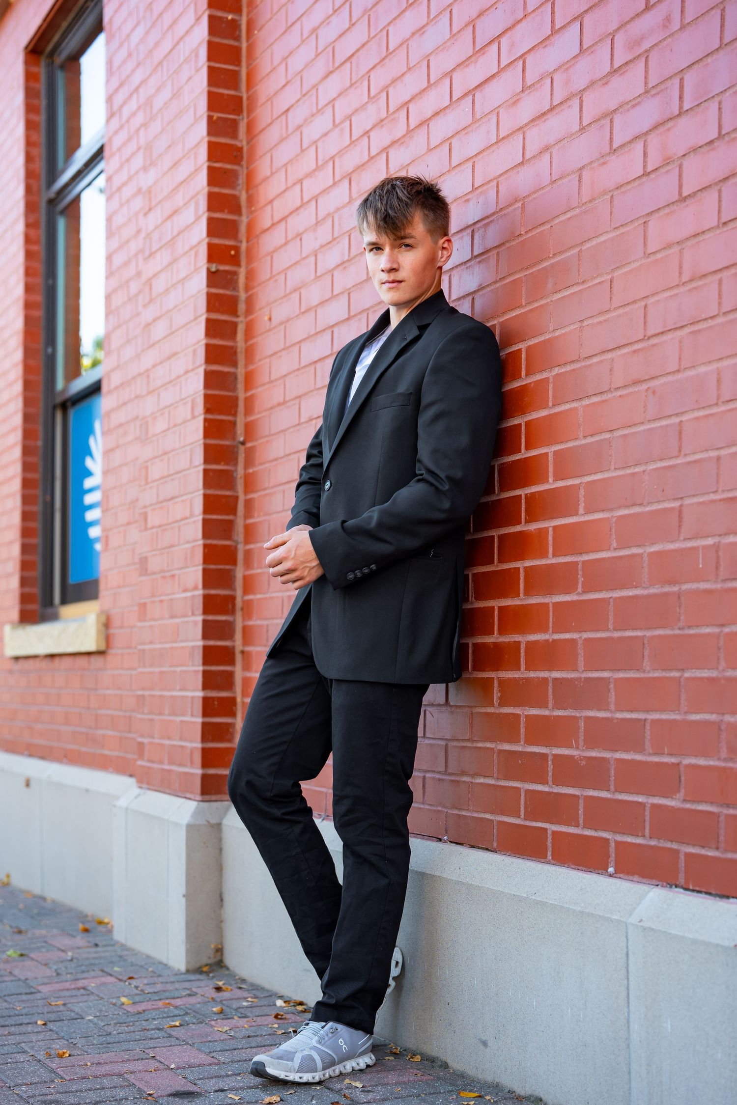 Person in formal black suit standing against pink brick wall with windows.