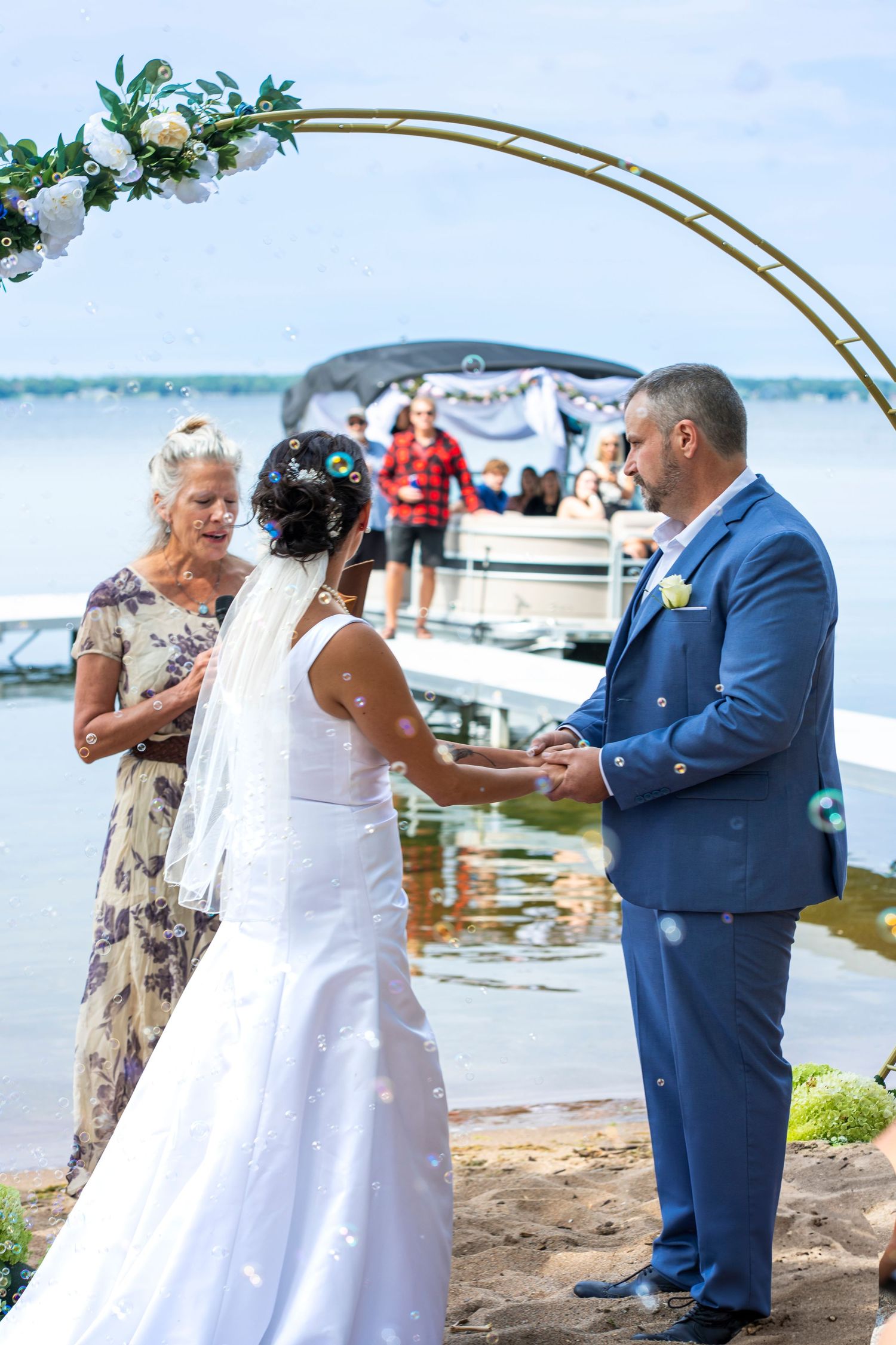 Wedding ceremony under copper arch by waterfront with boats in background.