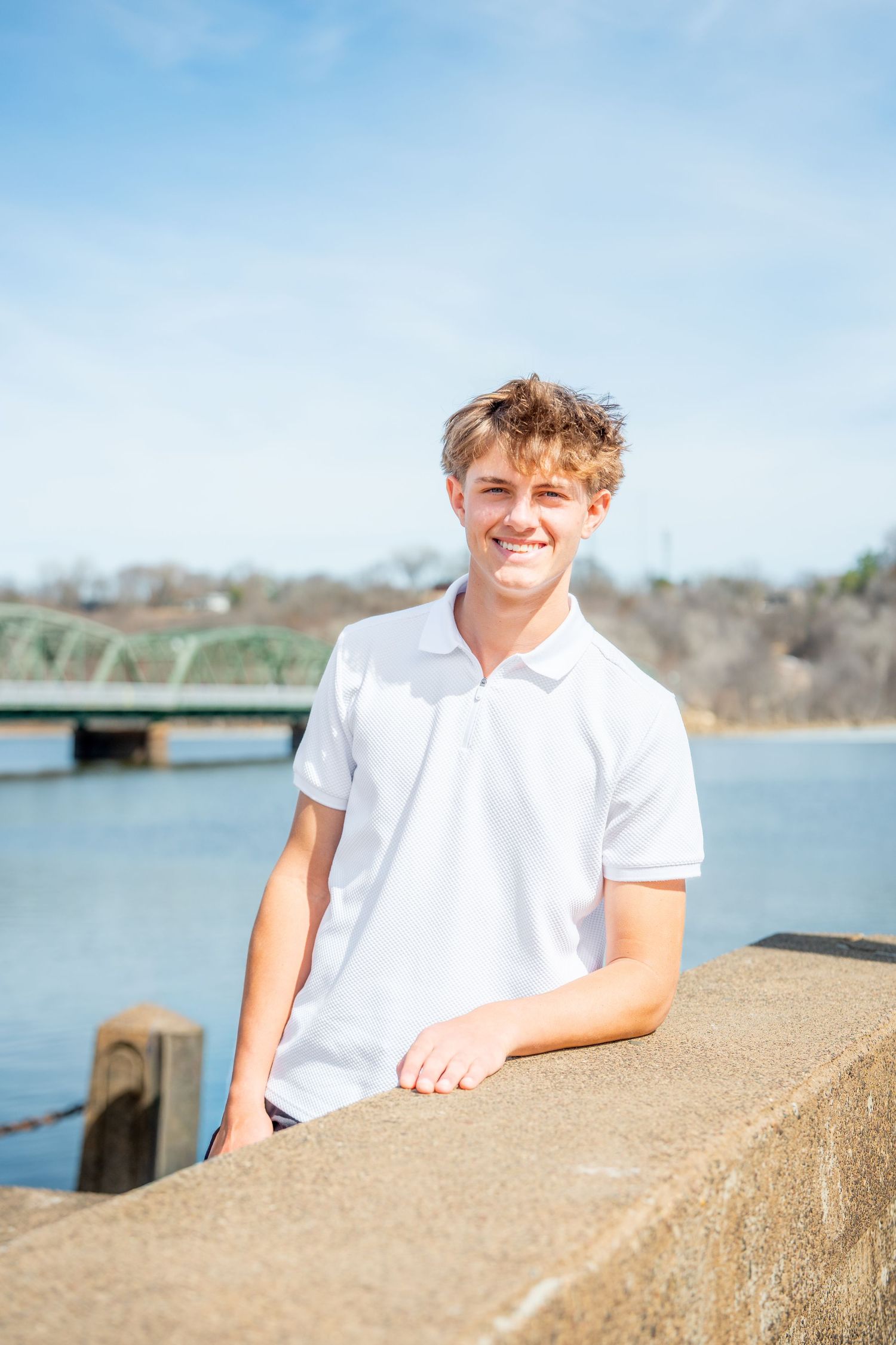 A person in a white polo shirt stands by a concrete wall near a bridge over blue water on a sunny day.