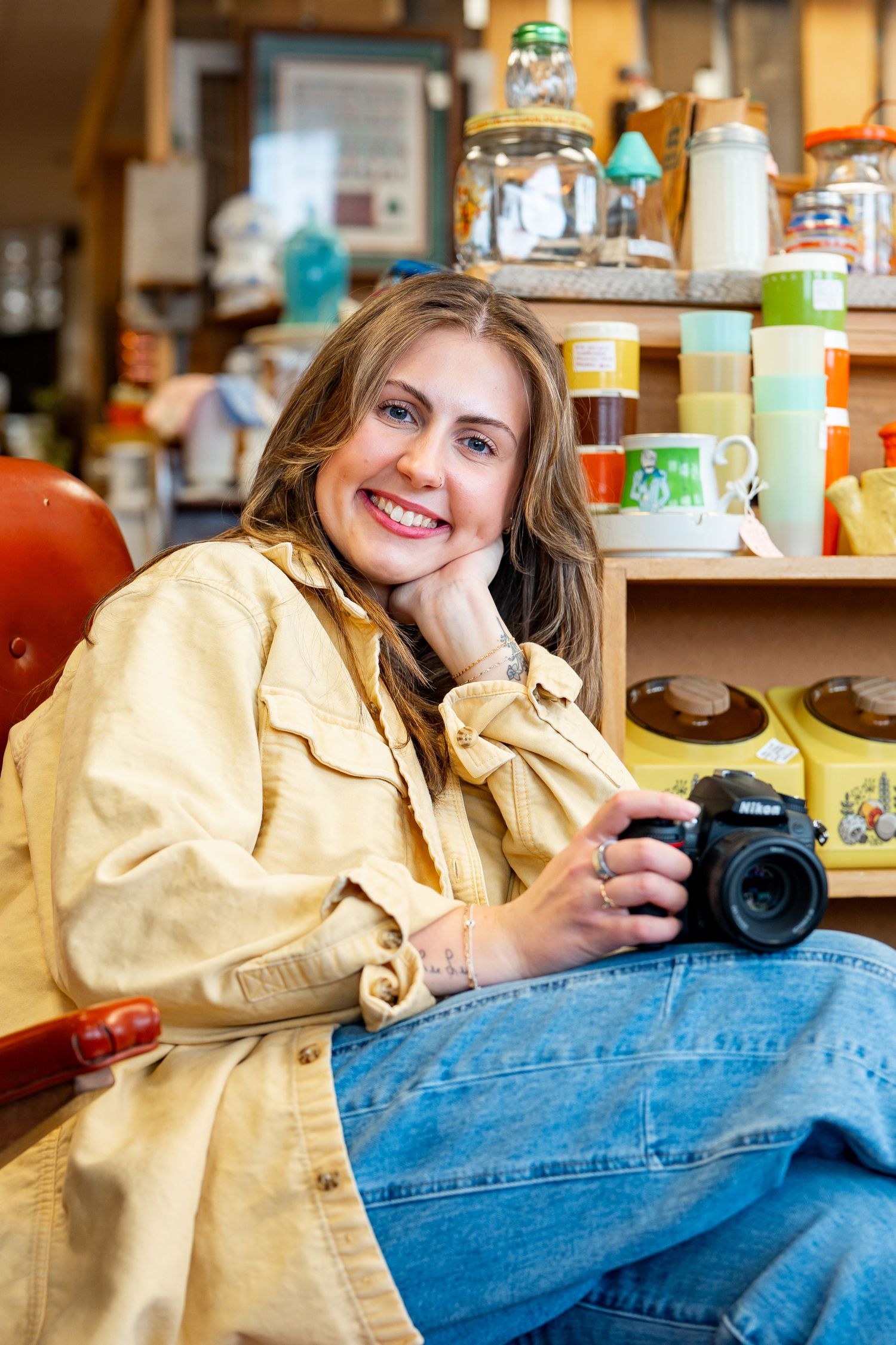 Smiling woman holding a Nikon camera sits in a vintage shop surrounded by colorful retro kitchenware and collectibles.