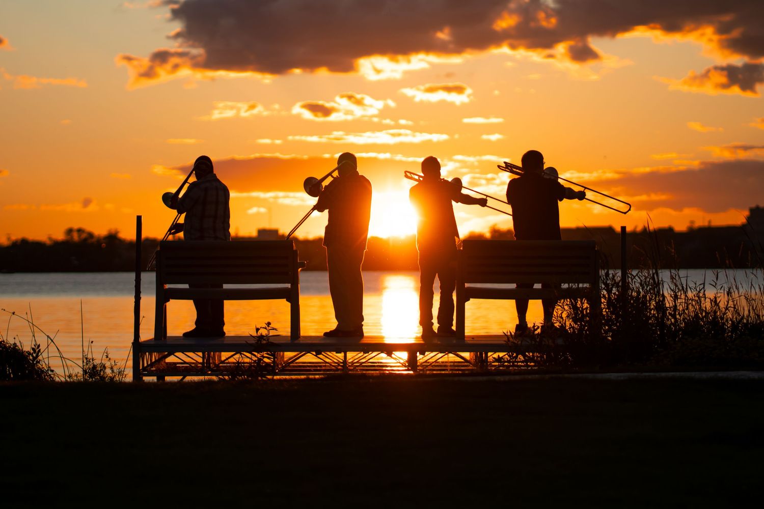 Silhouettes of people against dramatic orange sunset sky at waterfront playing trombone.