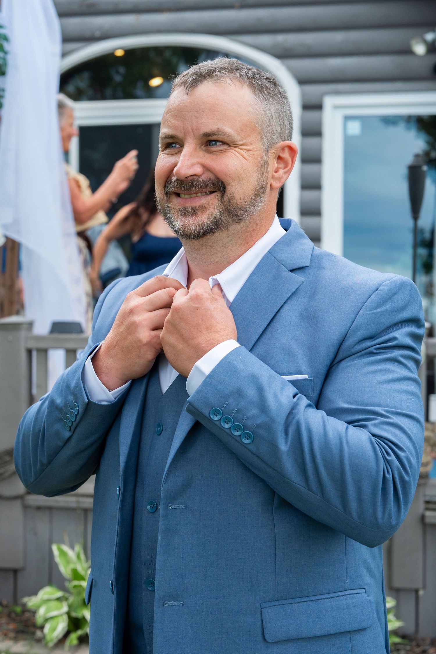 Groom in blue suit laughing and gesturing during outdoor wedding ceremony.