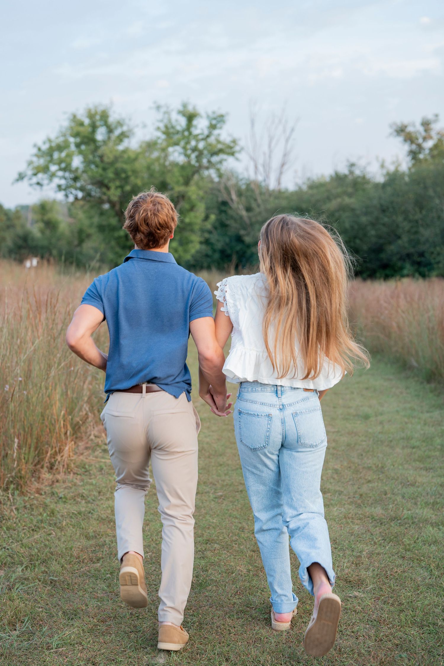 A couple walks away hand in hand through a grassy field on a sunny day.