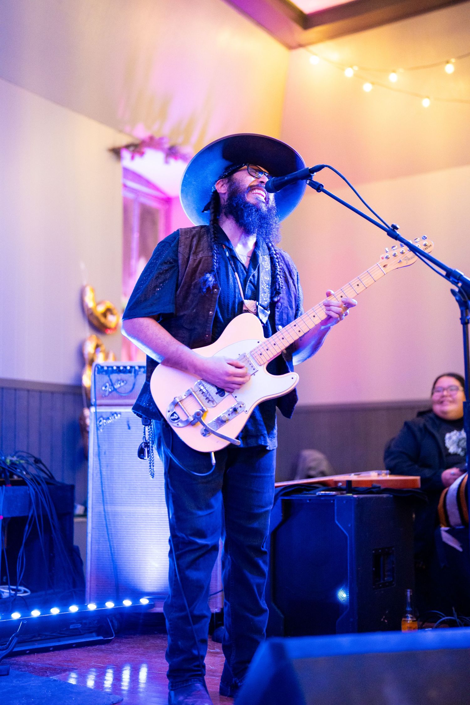 Musician in a wide-brimmed hat playing guitar on stage with string lights overhead.