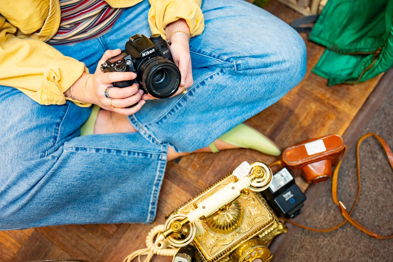 Person in yellow jacket sitting cross-legged examining a DSLR camera surrounded by vintage cameras on wooden floor.
