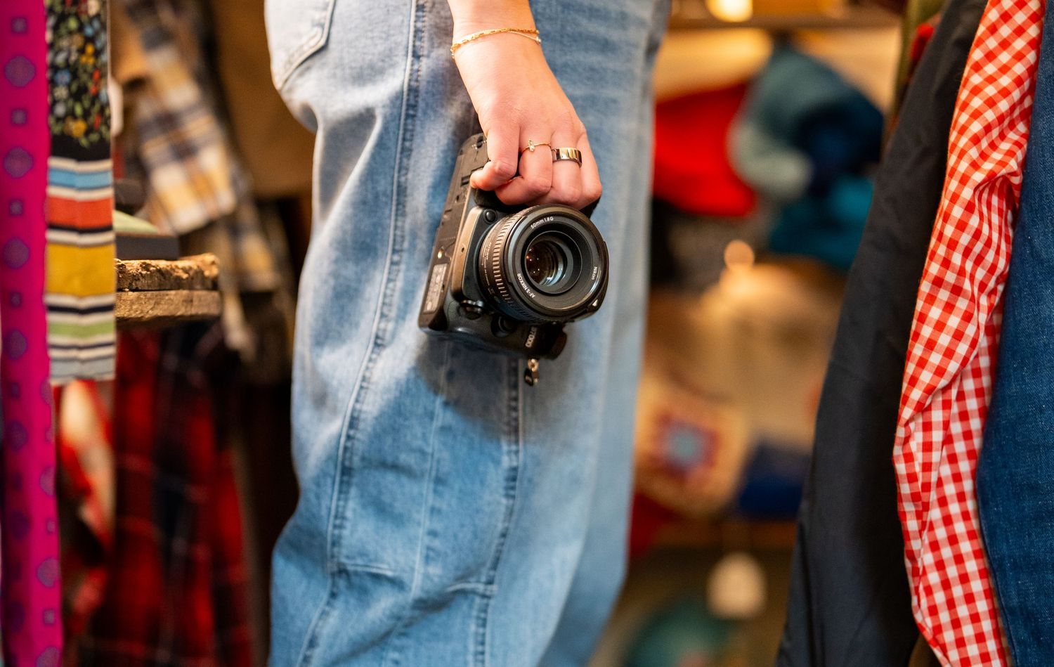 Person holding a black camera while browsing colorful clothing racks in a vintage fashion boutique.