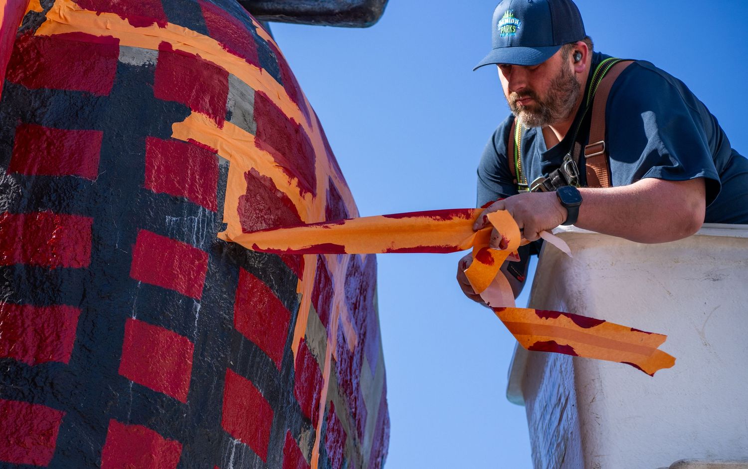 Artist painting details on a large Paul Bunyan statue wearing a red plaid shirt against a bright blue sky.
