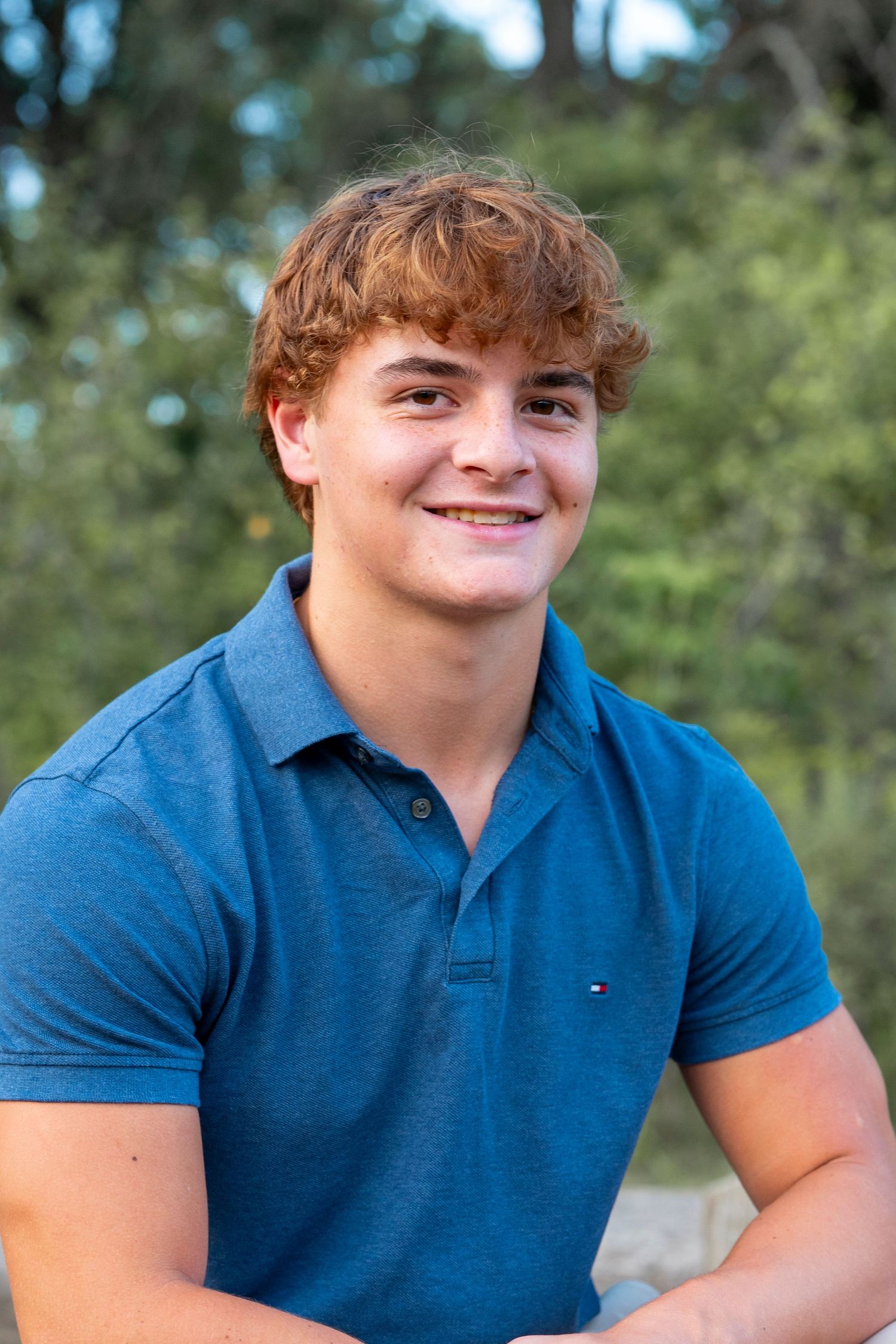 Senior portrait in nature wearing a blue polo shirt against a blurred forest background.