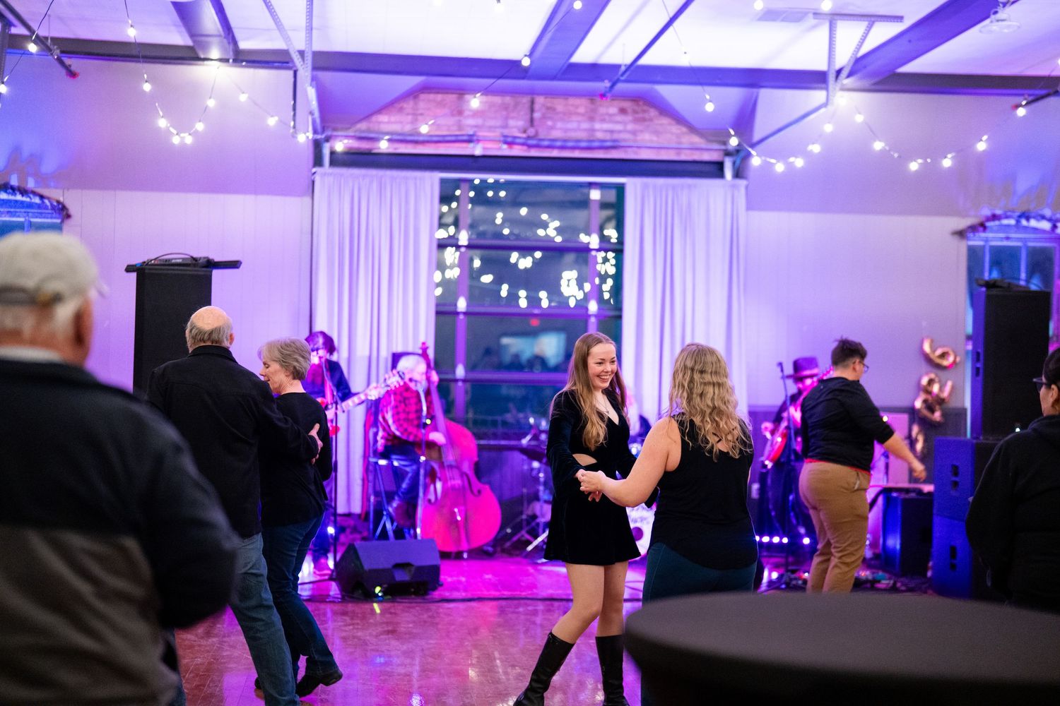 People dancing at an indoor event venue with purple ambient lighting and string lights.