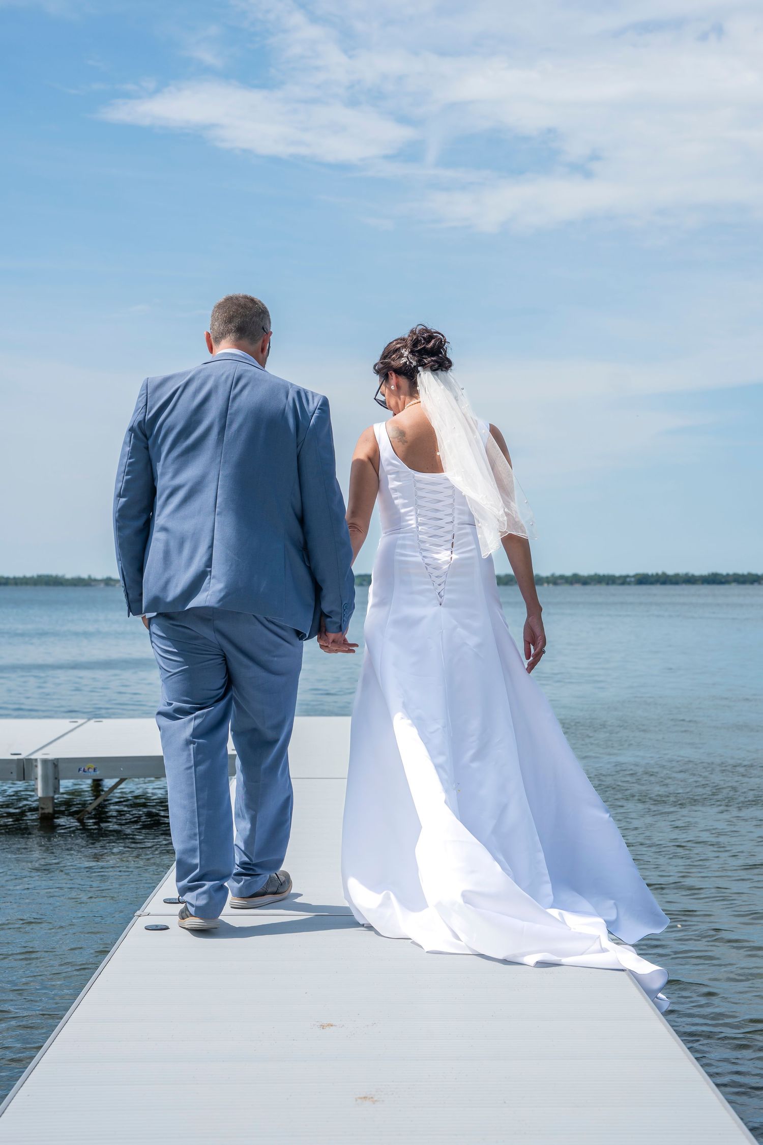 A couple in wedding attire sits together on a white dock overlooking a serene lake on a sunny day.