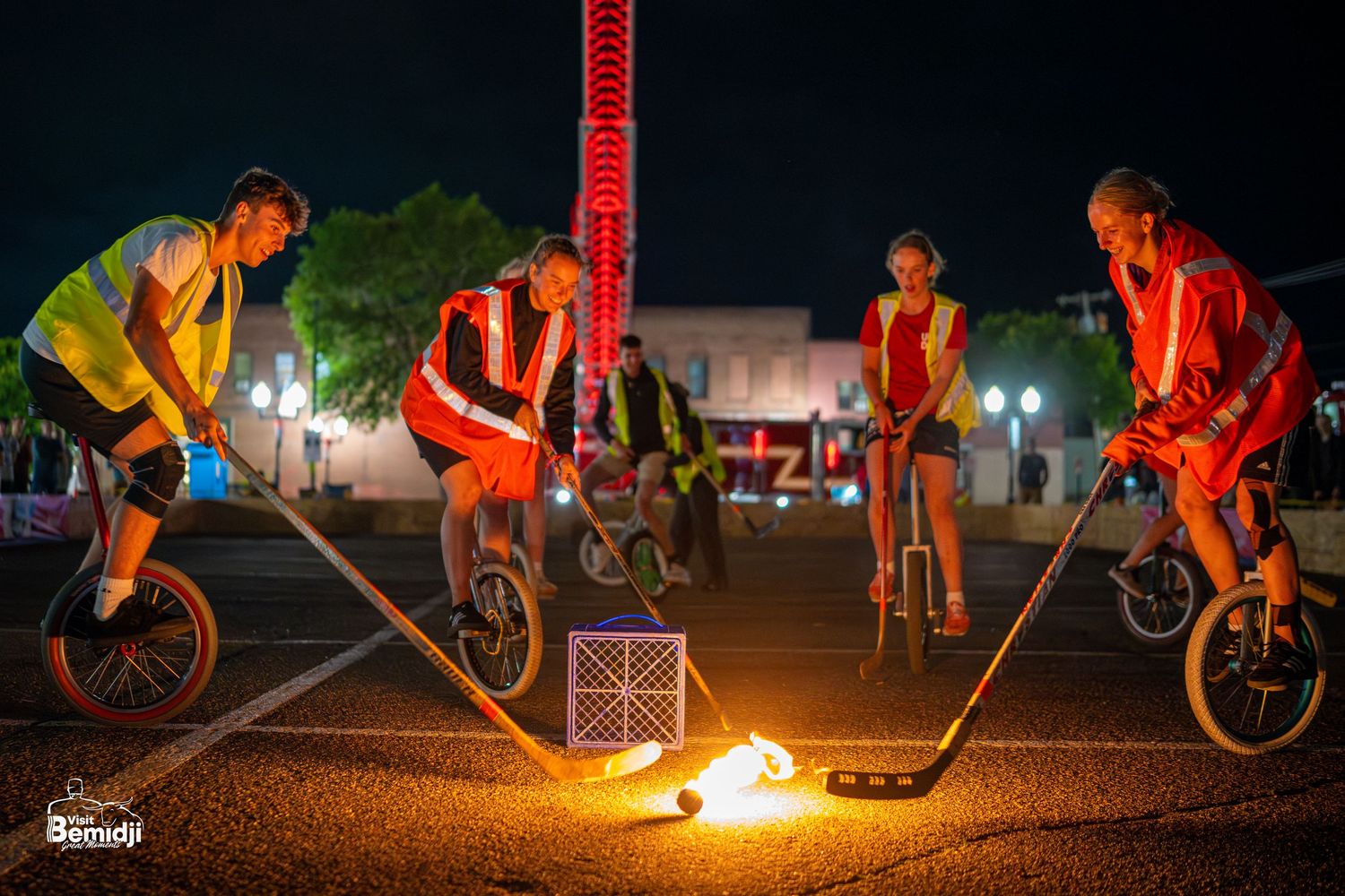 Unicyclists playing fire hockey at night on an illuminated court.