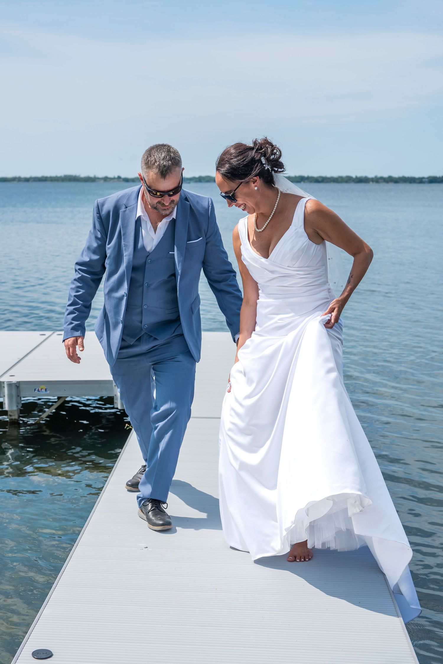 Newlyweds in formal attire share lighthearted moment on dock by peaceful lake.