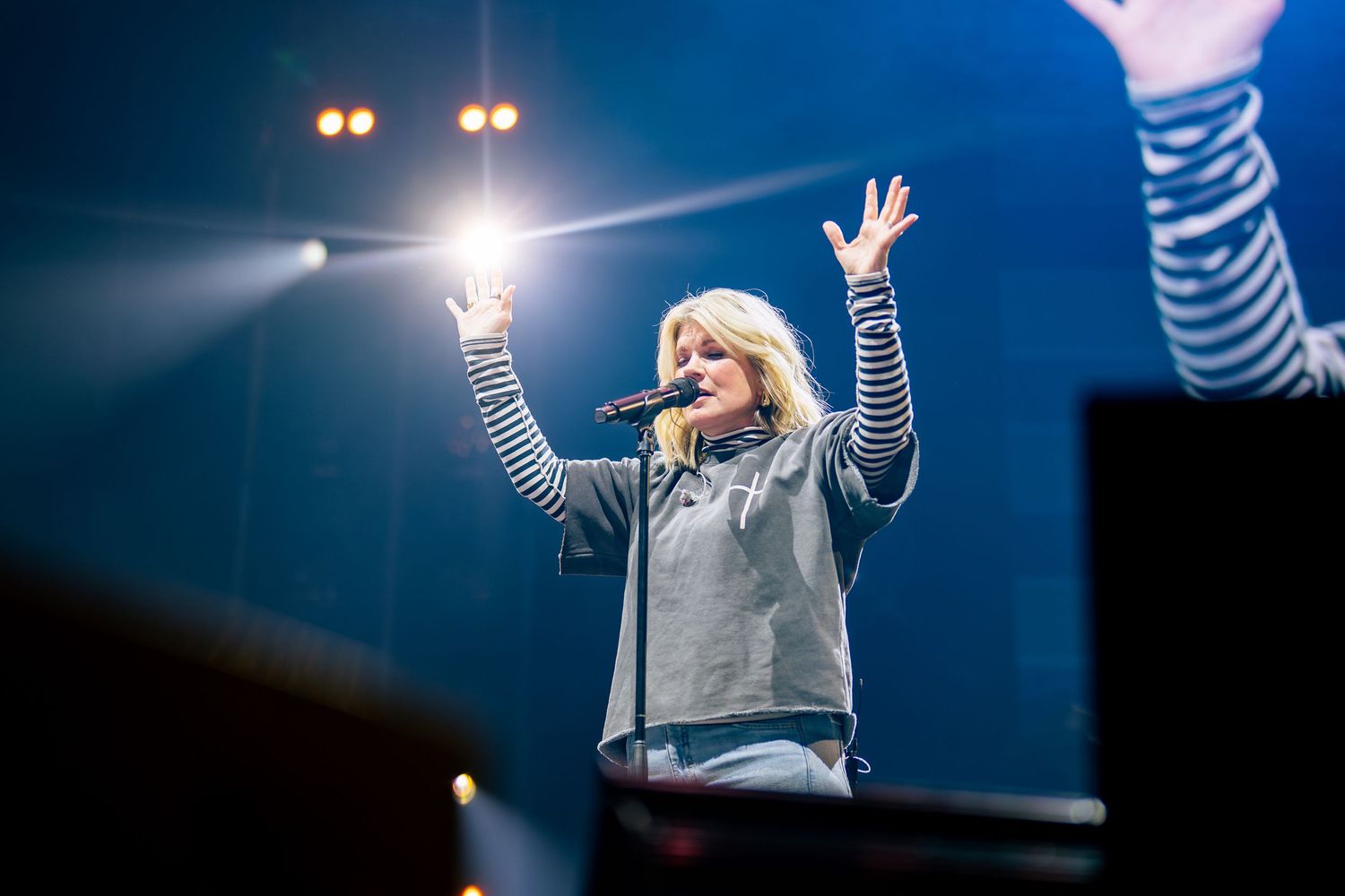 A concert performer in a gray top raises their arms during an energetic stage performance with dramatic lighting.