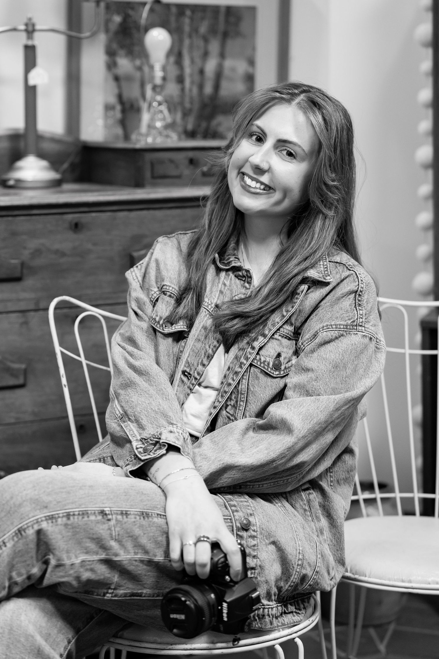 Smiling woman in denim jacket sits on wire chair holding camera in black and white portrait photo.