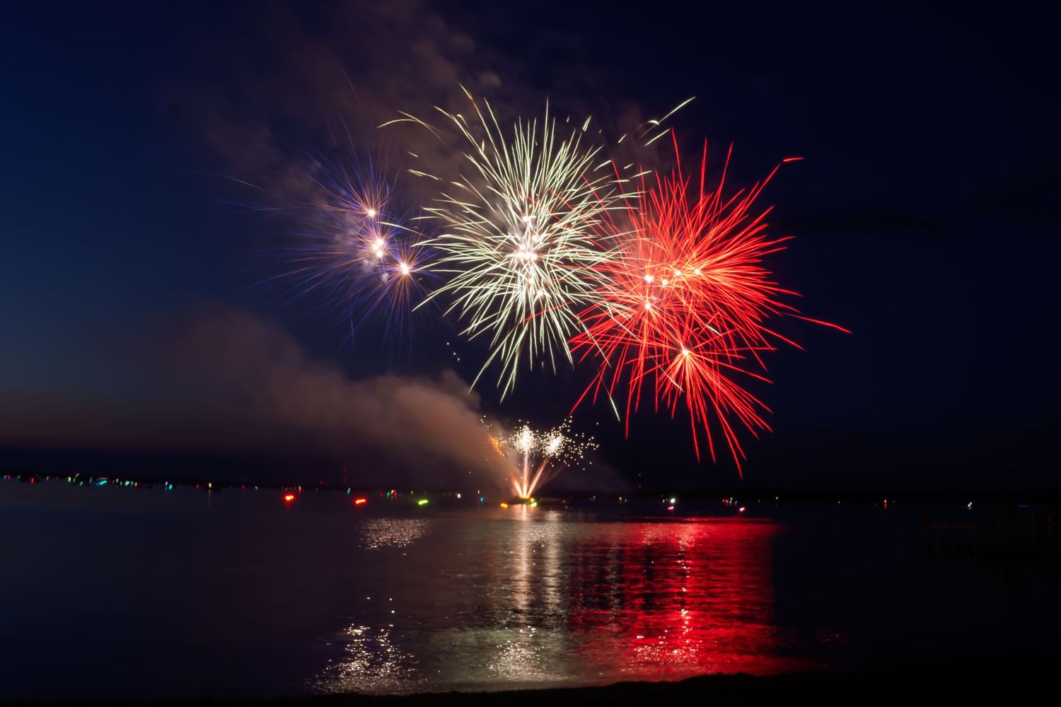 Red and white fireworks exploding over water at night with reflections on the surface.